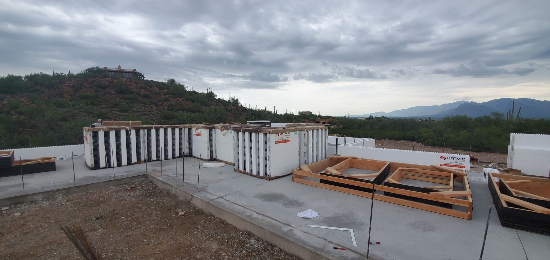 Construction site with insulated concrete forms, surrounded by vegetation, under a cloudy sky.