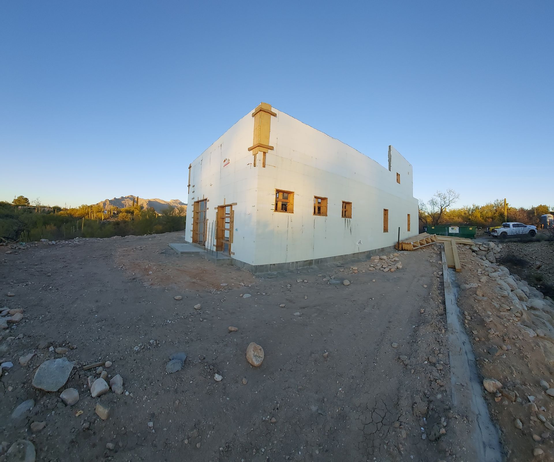 White building under construction, brown wooden doors/window frames, dirt ground, blue sky.