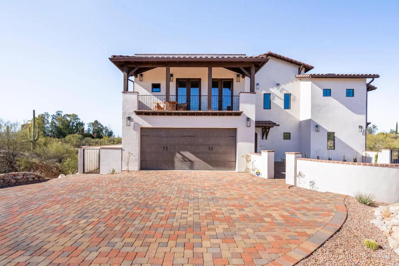Two-story white house with brick driveway; balcony on top floor, brown garage door, against a blue sky.
