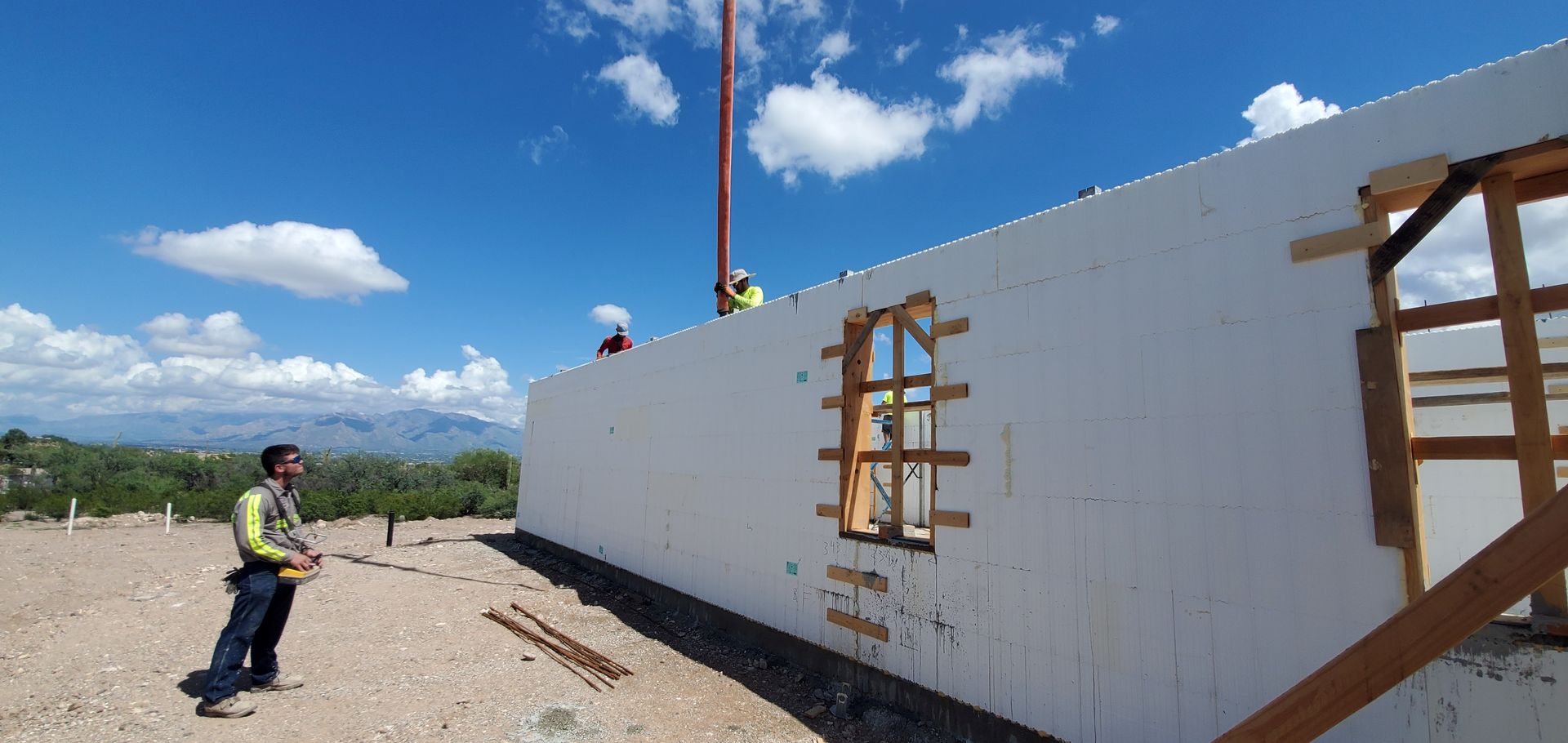 Construction workers near a building. One holds tools, the others on roof near a pole. Blue sky, mountains in the distance.