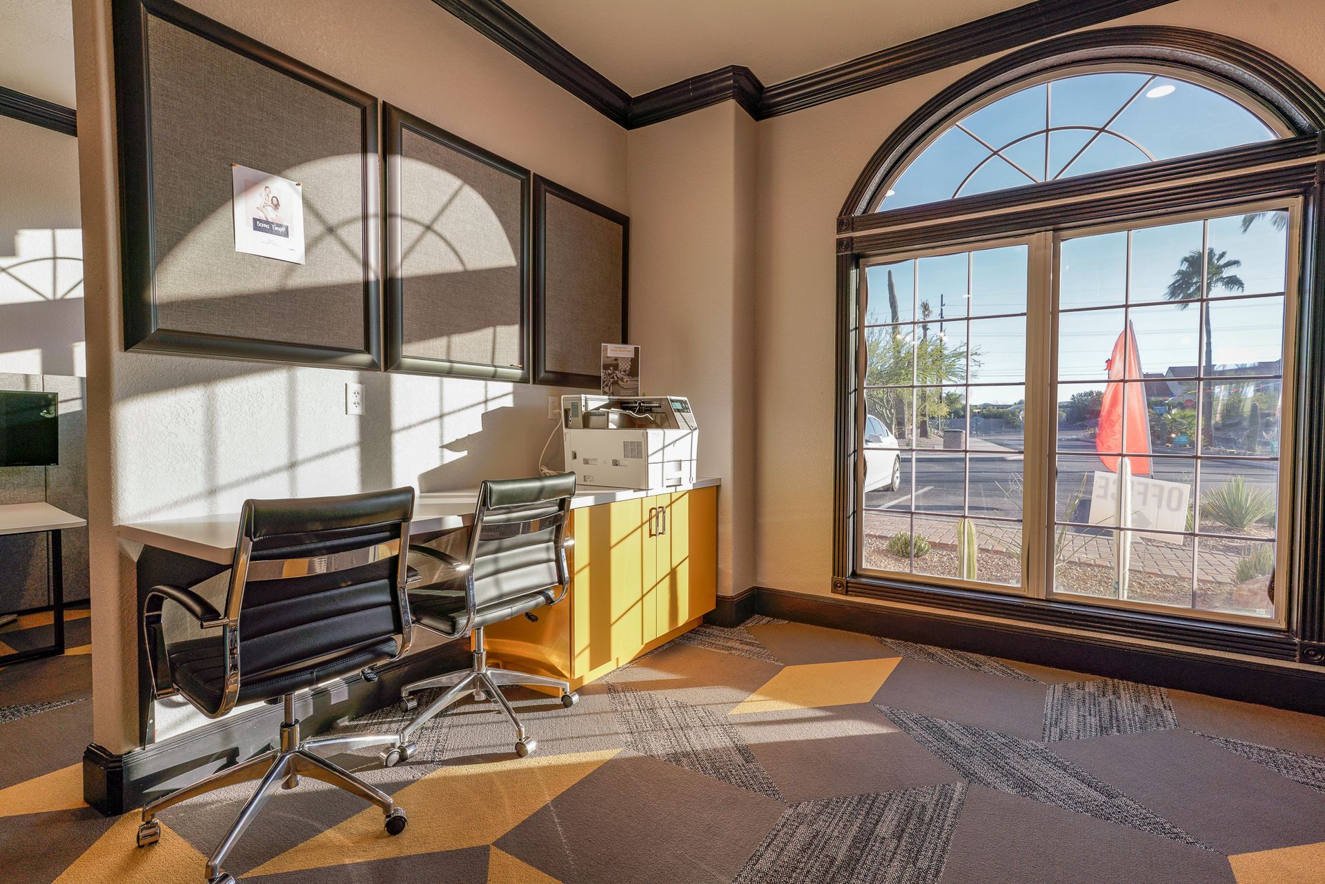 Office with large arched window, chairs, corkboards, and printer, with sunlight shining in.