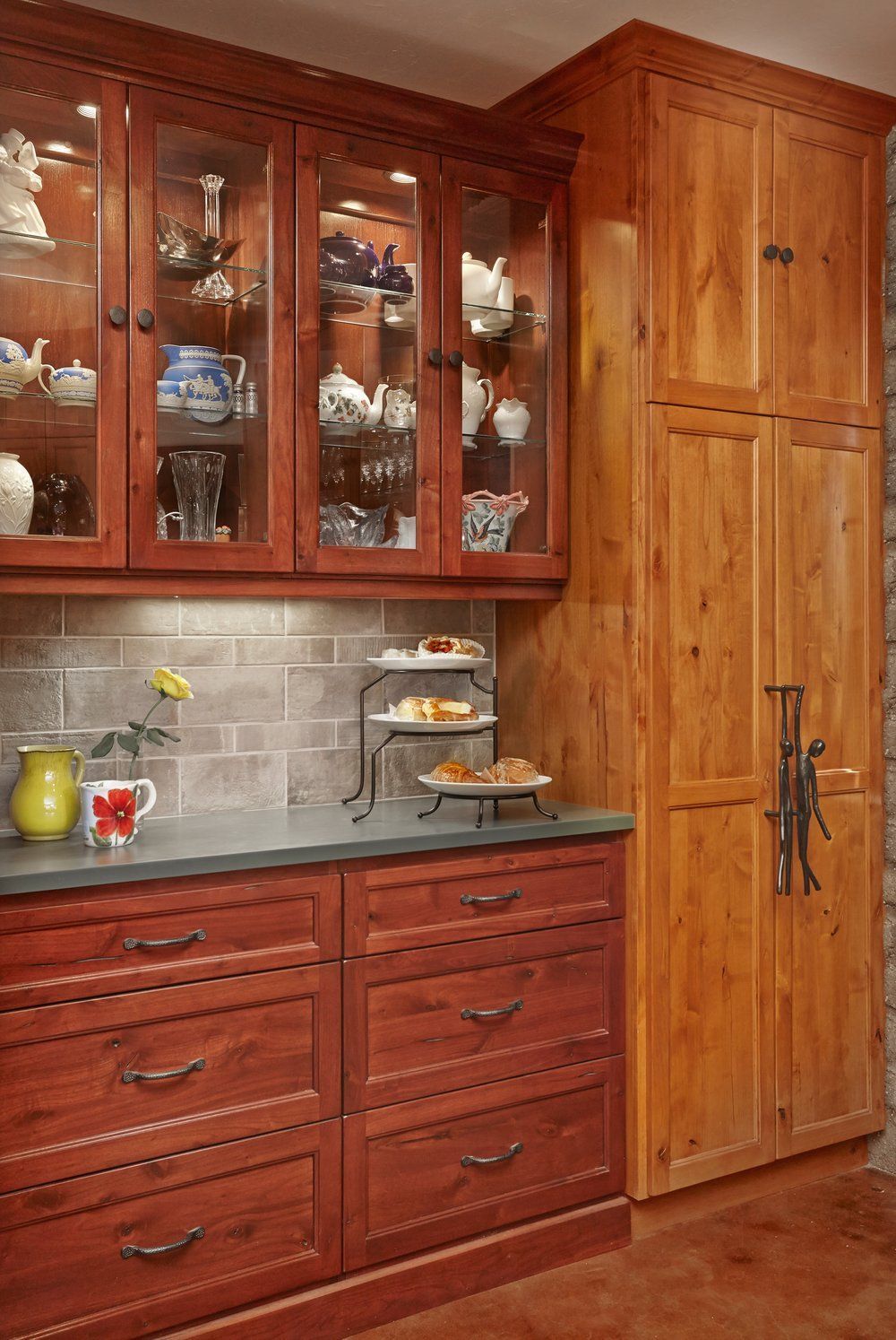 Kitchen cabinetry: glass-fronted display, drawers, and tall pantry with brown-red wood and a stone backsplash.