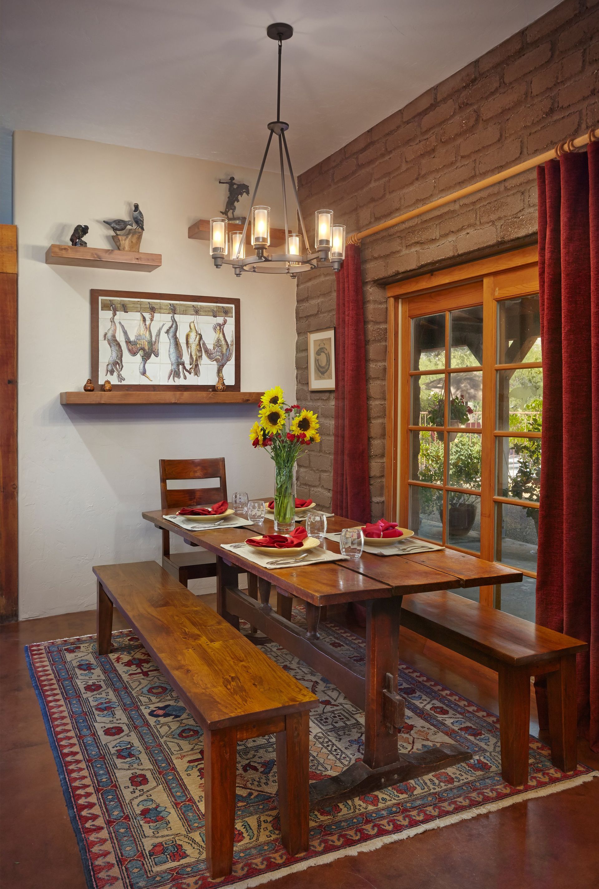 Dining room with wooden table, benches, and a colorful rug under a chandelier; brick wall and sunflowers.