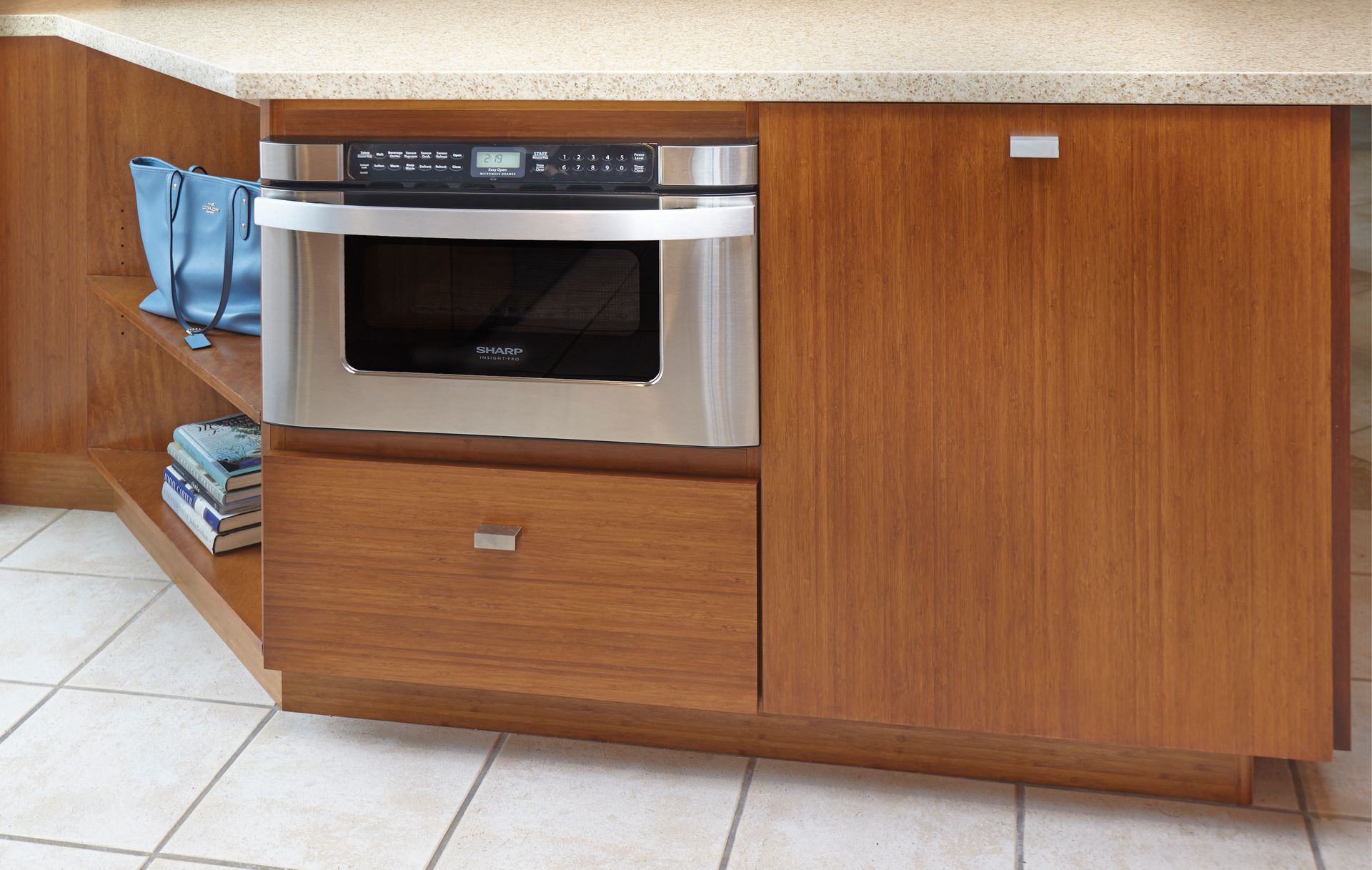 Built-in microwave oven and drawer in wooden kitchen cabinetry; blue bag and books on open shelf.