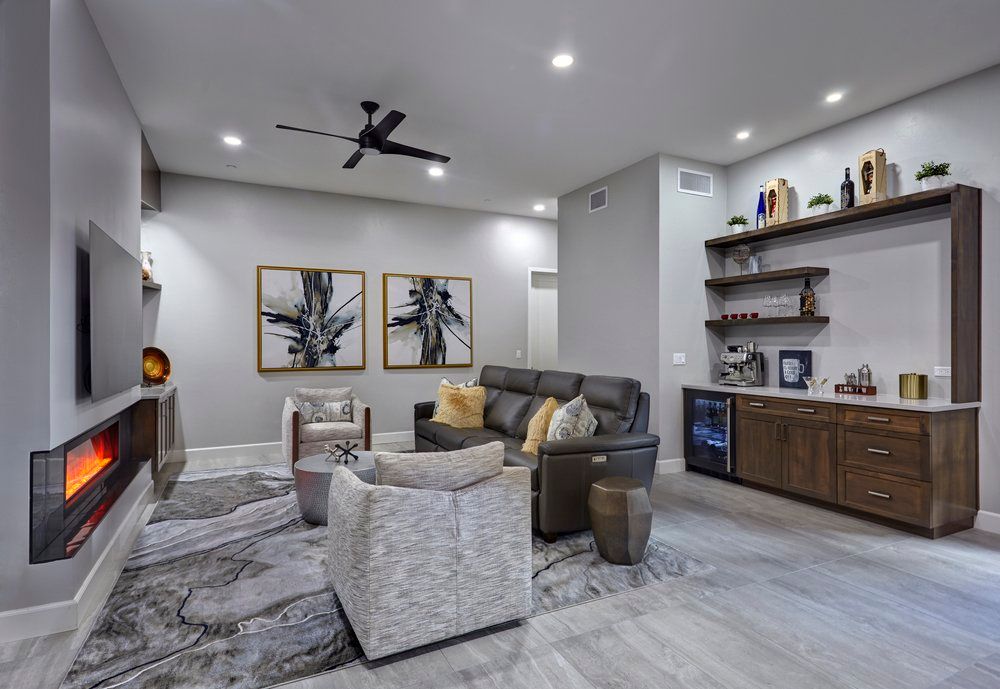Modern living room with fireplace, gray walls, bar, gray and brown furniture, artwork, and ceiling fan.