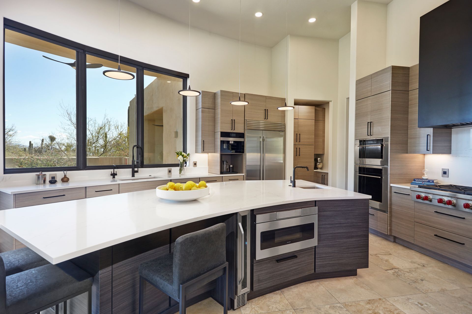 Modern kitchen with island, stainless steel appliances, large windows, light wood cabinets, white countertops.