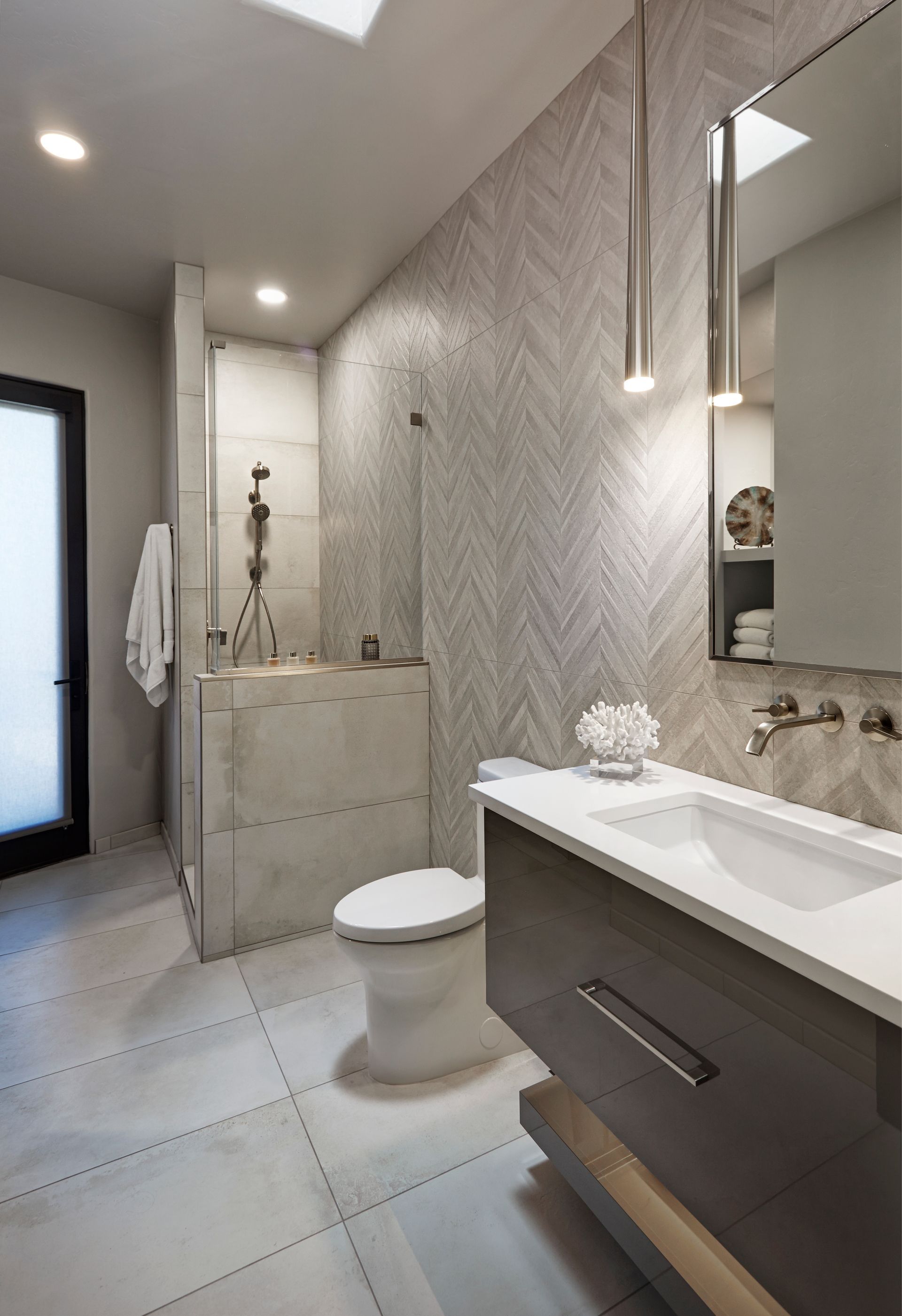 Modern bathroom with shower, toilet, and vanity. Gray and white color scheme, herringbone accent wall, and natural light.