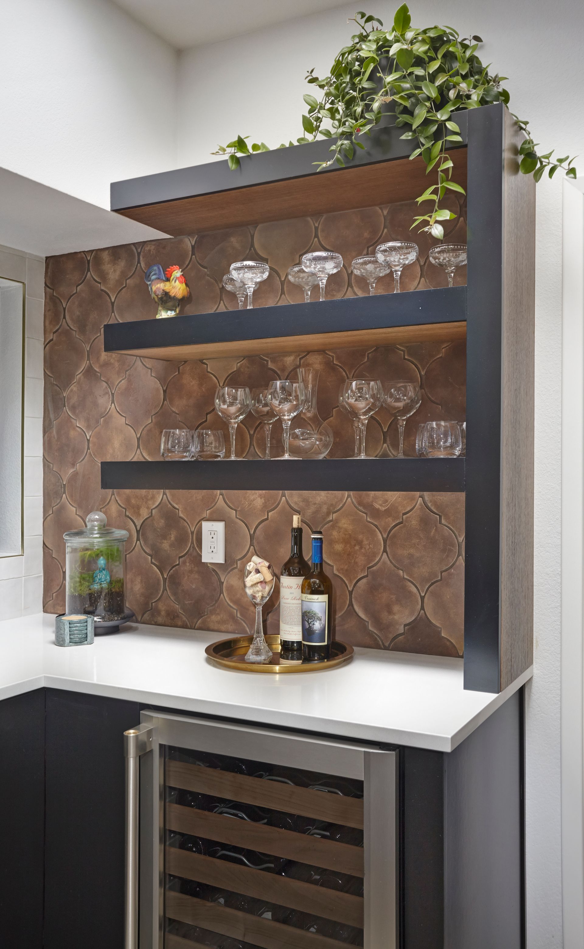 Bar area with tiered shelves displaying glassware. Wine bottles, tray, and a plant. Brown and black accents.