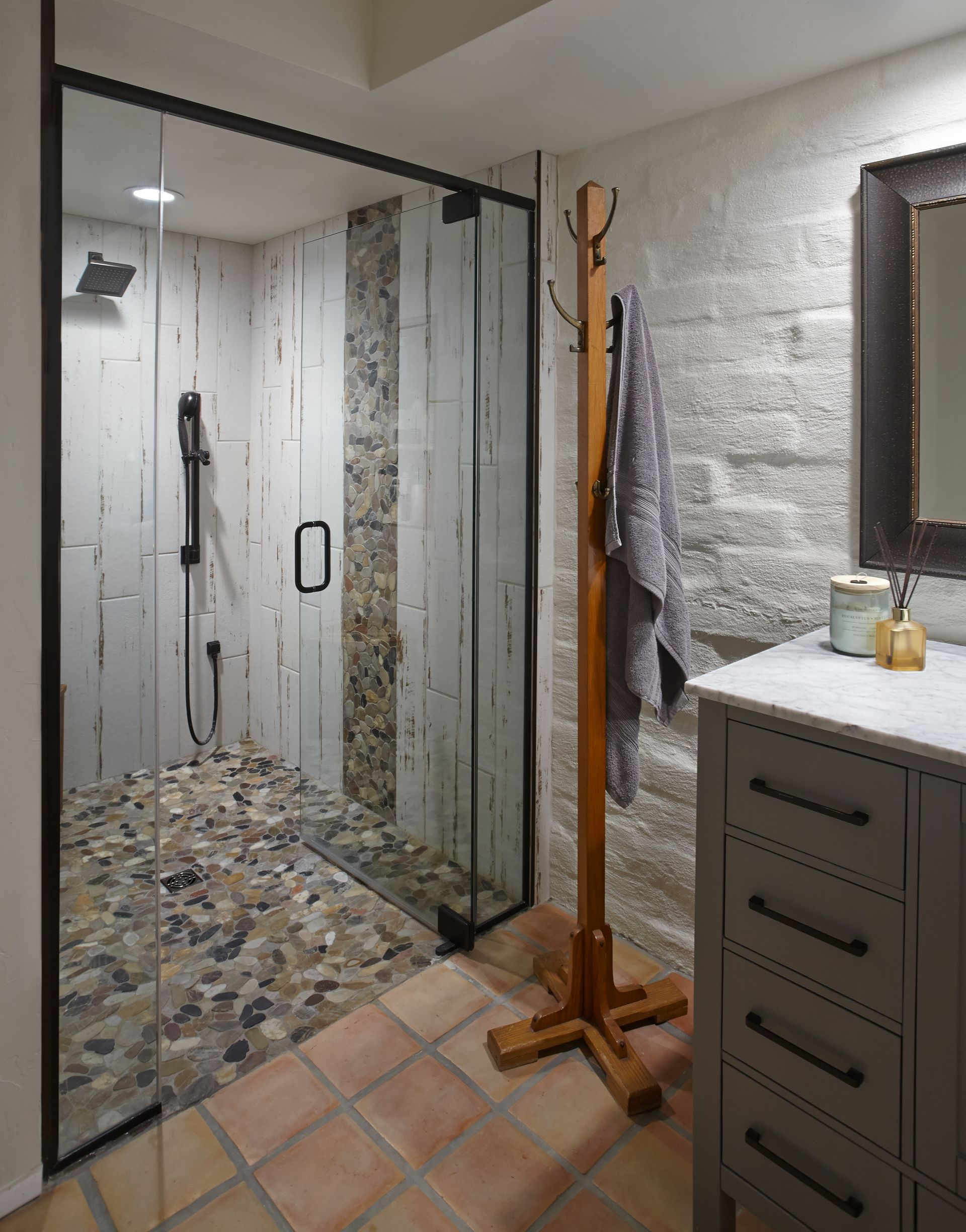 Shower with glass door, stone floor, pebble shower floor, wooden coat rack, grey vanity, stone wall.