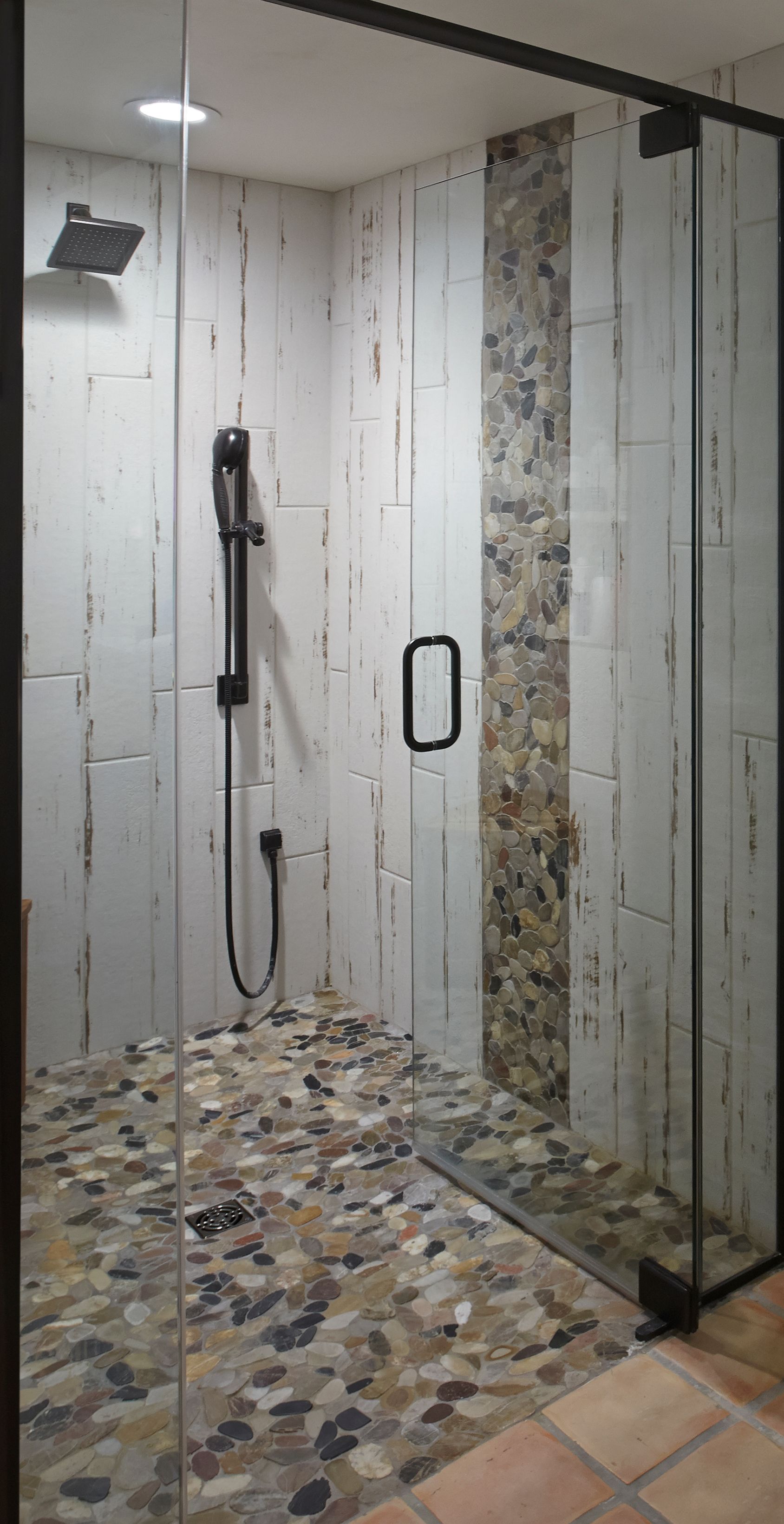 Shower with glass door, pebble floor, white vertical tiles, black fixtures, and stone accent.