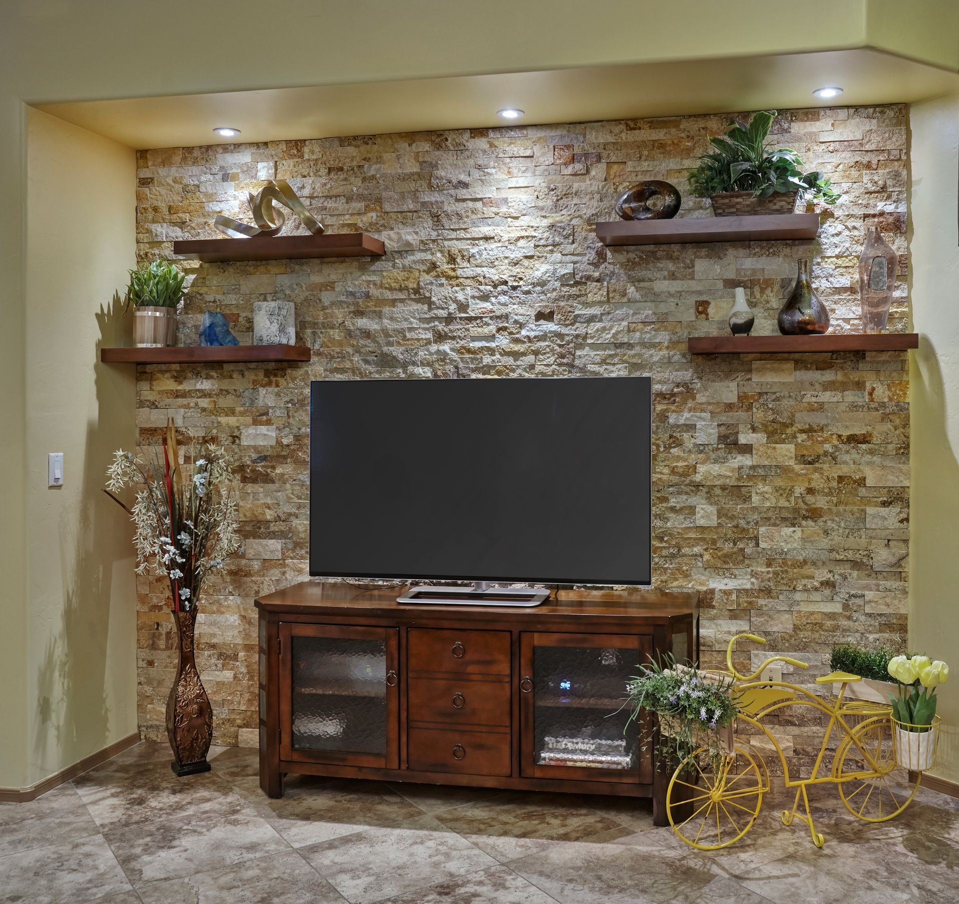 Living room with stone accent wall, TV, wooden shelves with decor, and brown cabinet.