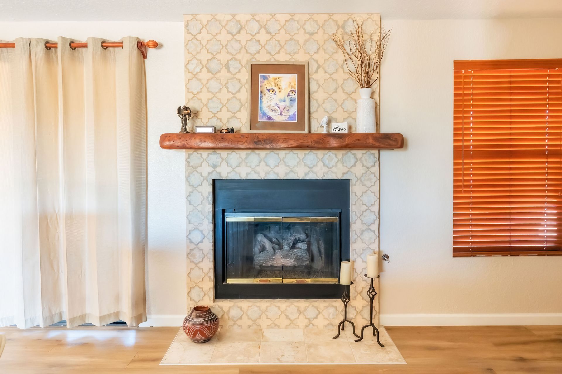 Fireplace with patterned tile surround, wooden mantle, and window with orange blinds.