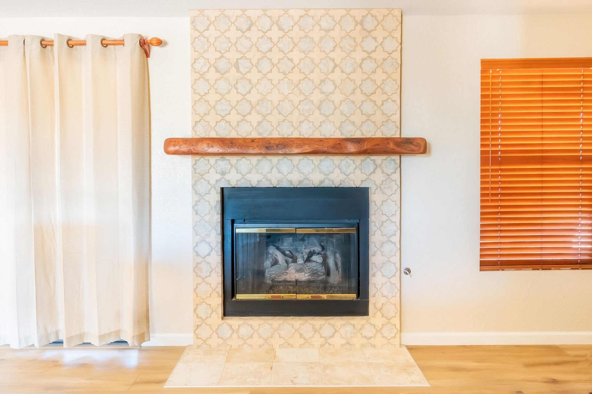 Fireplace with patterned tile surround, wooden mantel, and wooden blinds. Light beige curtains.