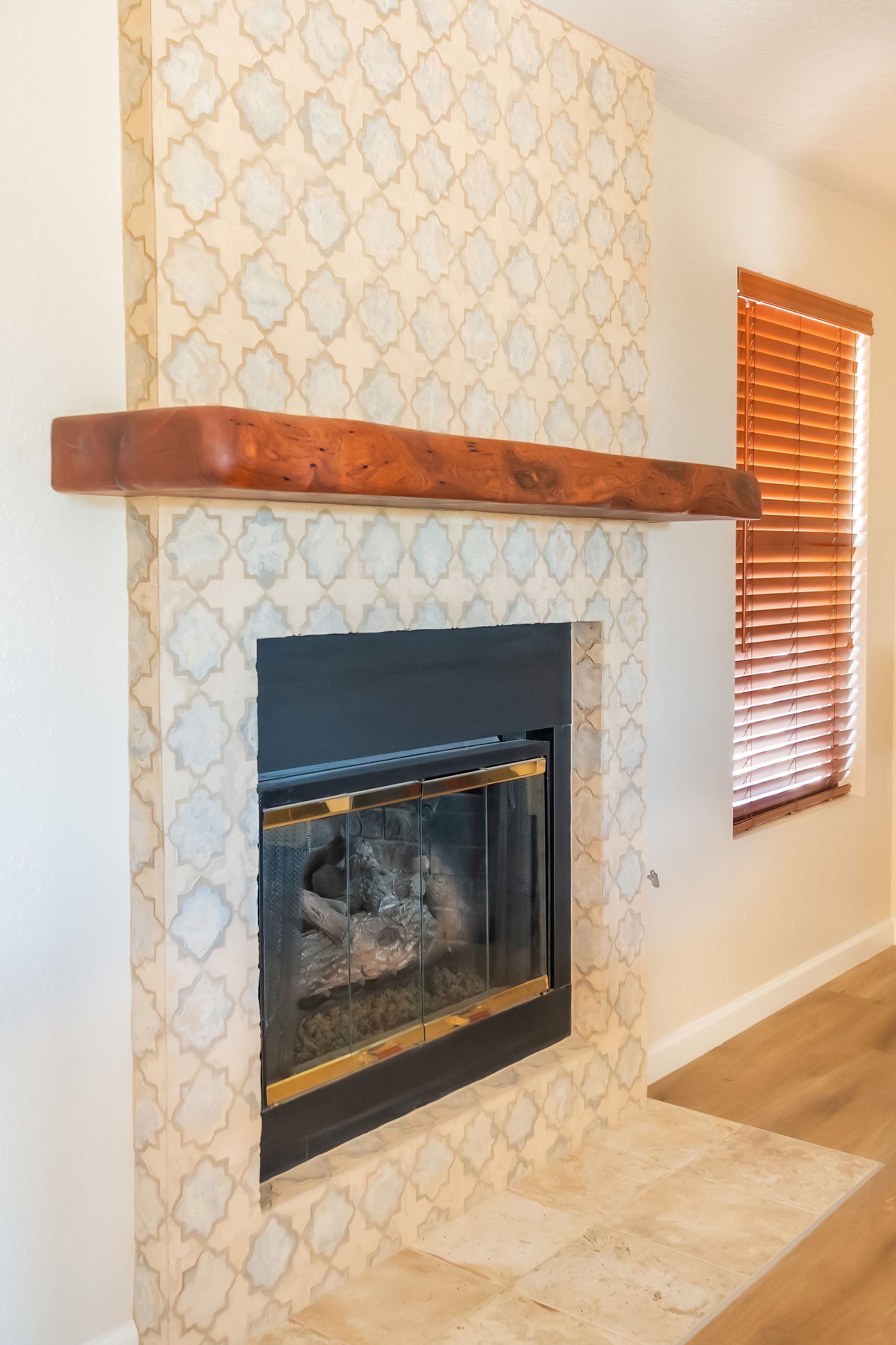 Fireplace with ornate tile surround, wooden mantel, and wooden blinds on a window.