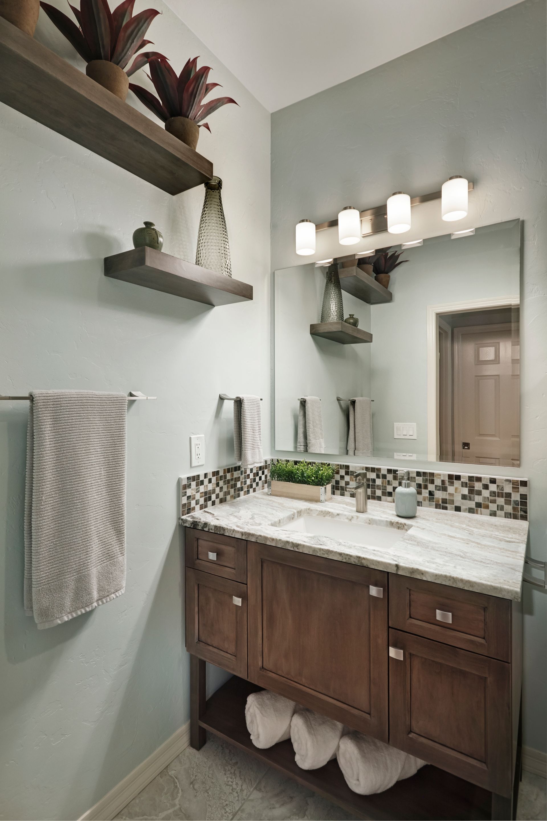 Bathroom with wooden vanity, shelves, and mirror. Gray-blue walls, white countertop, and mosaic tile backsplash.