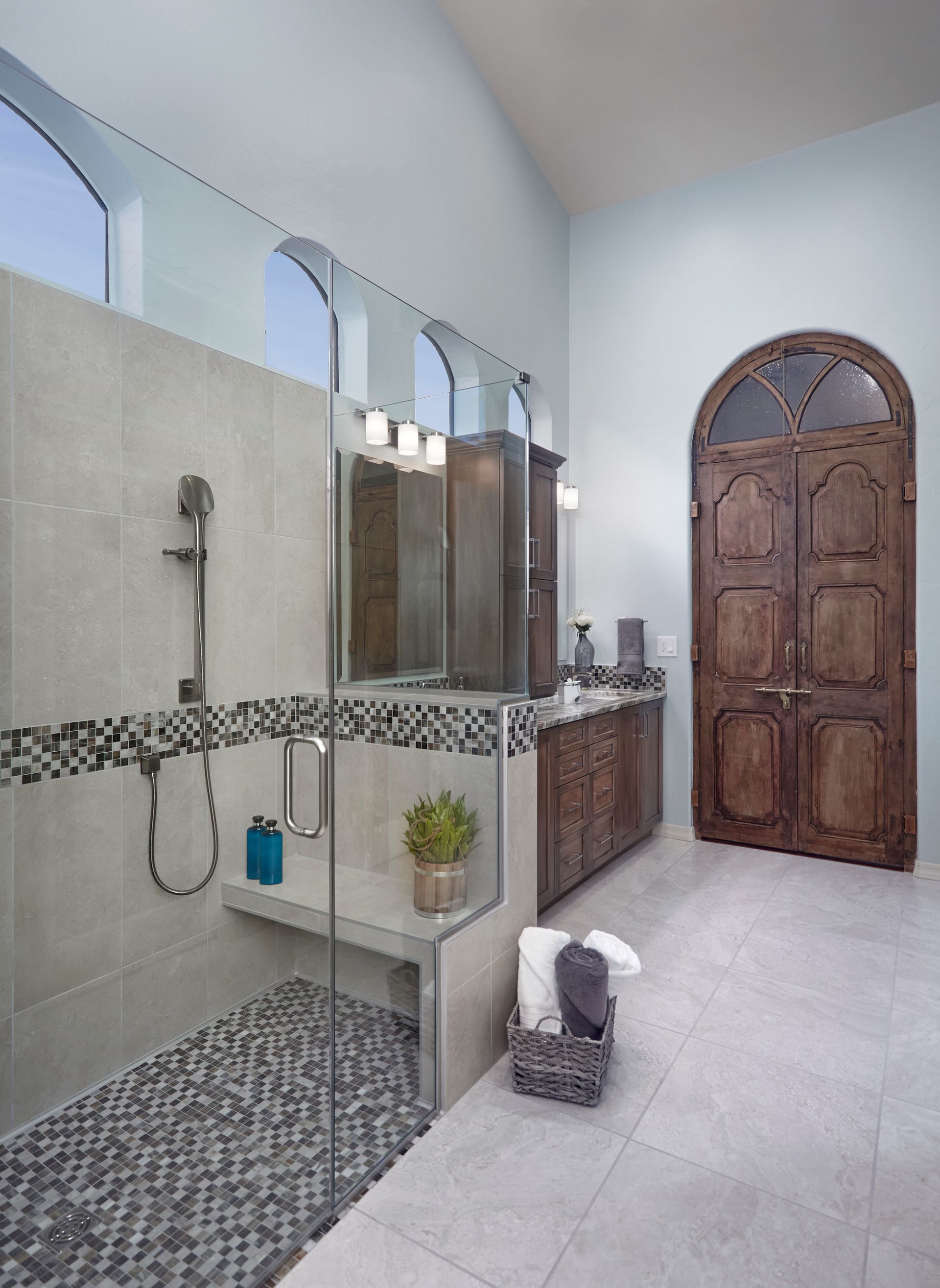 Spacious bathroom with glass shower, wooden cabinet, and arched wooden door; light blue walls, gray tile.