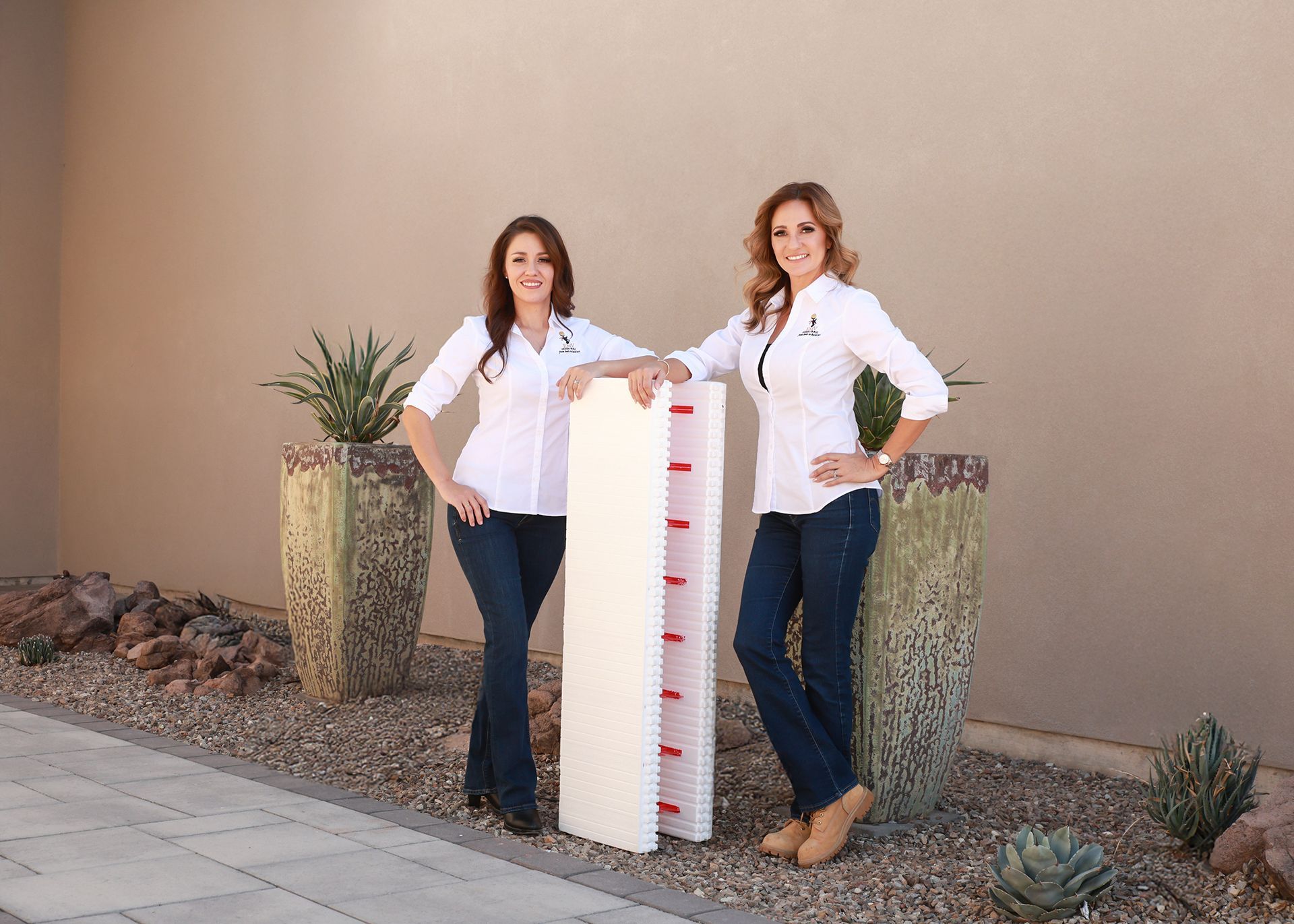 Two women stand by a large height measuring board outside. They wear white shirts, blue jeans, and smile.
