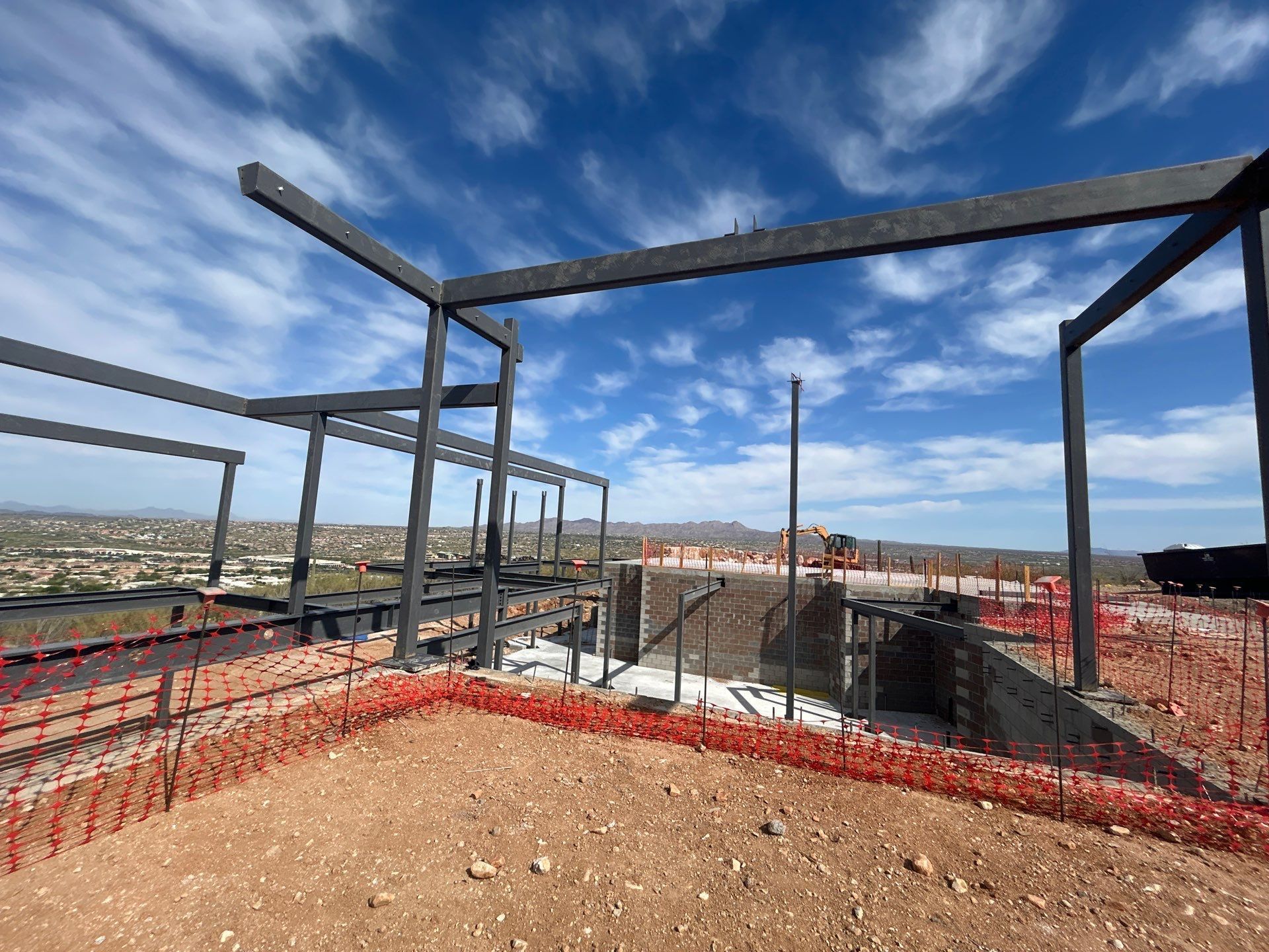 Construction site with black steel framework against a blue sky, surrounded by orange safety fencing.