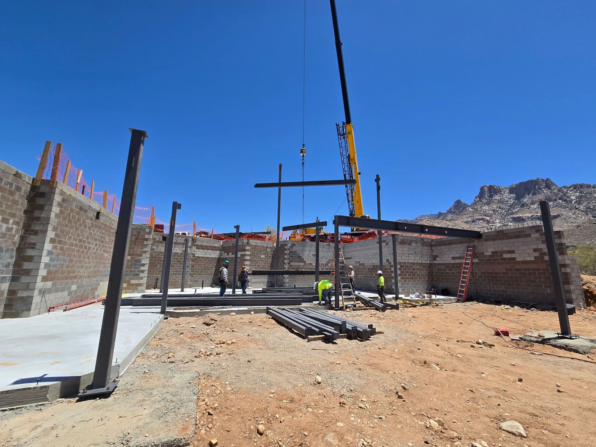 Construction site with crane lifting steel beams; workers on site, brick walls, concrete base, sunny day.