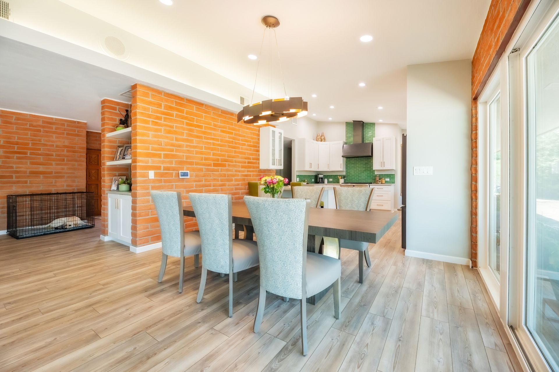 Dining room with brick accent wall, light wood floor, and table with chairs. Kitchen visible in the background.