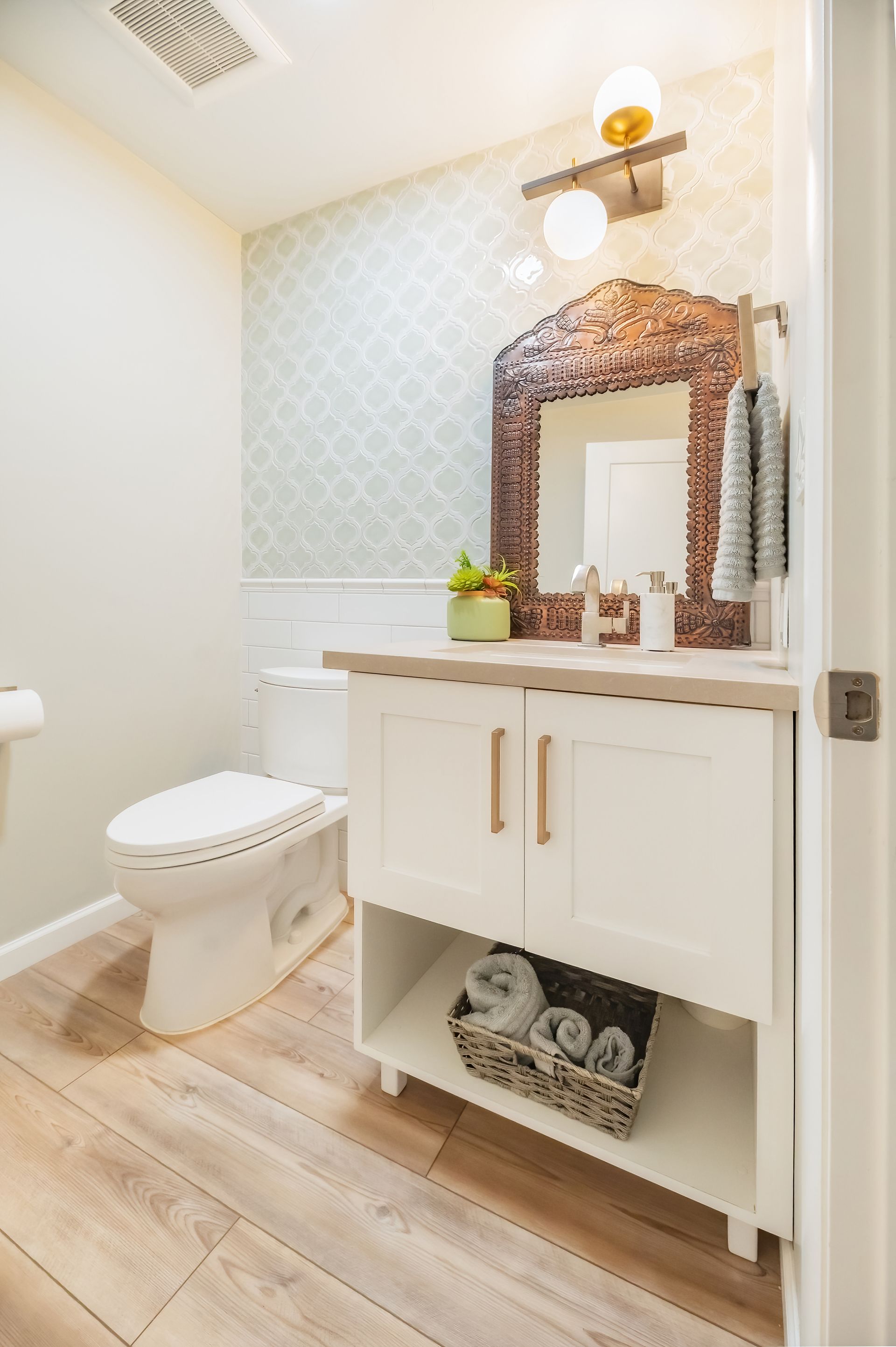 Bathroom with white vanity, toilet, and light wood flooring. Copper-framed mirror and patterned wallpaper.