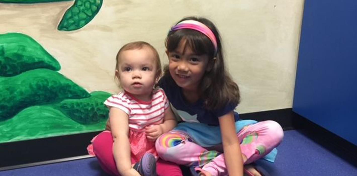 A young girl and baby in colorful clothes are sitting in a pediatric office in Peoria, AZ with a colorful wall. 