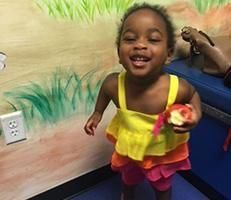A little girl is standing in a room holding an apple and smiling.
