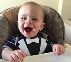 A baby wearing a tuxedo bib and bow tie is sitting in a high chair.