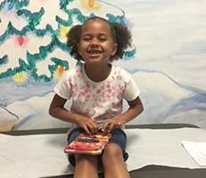A little girl is sitting on a table holding a book and smiling.