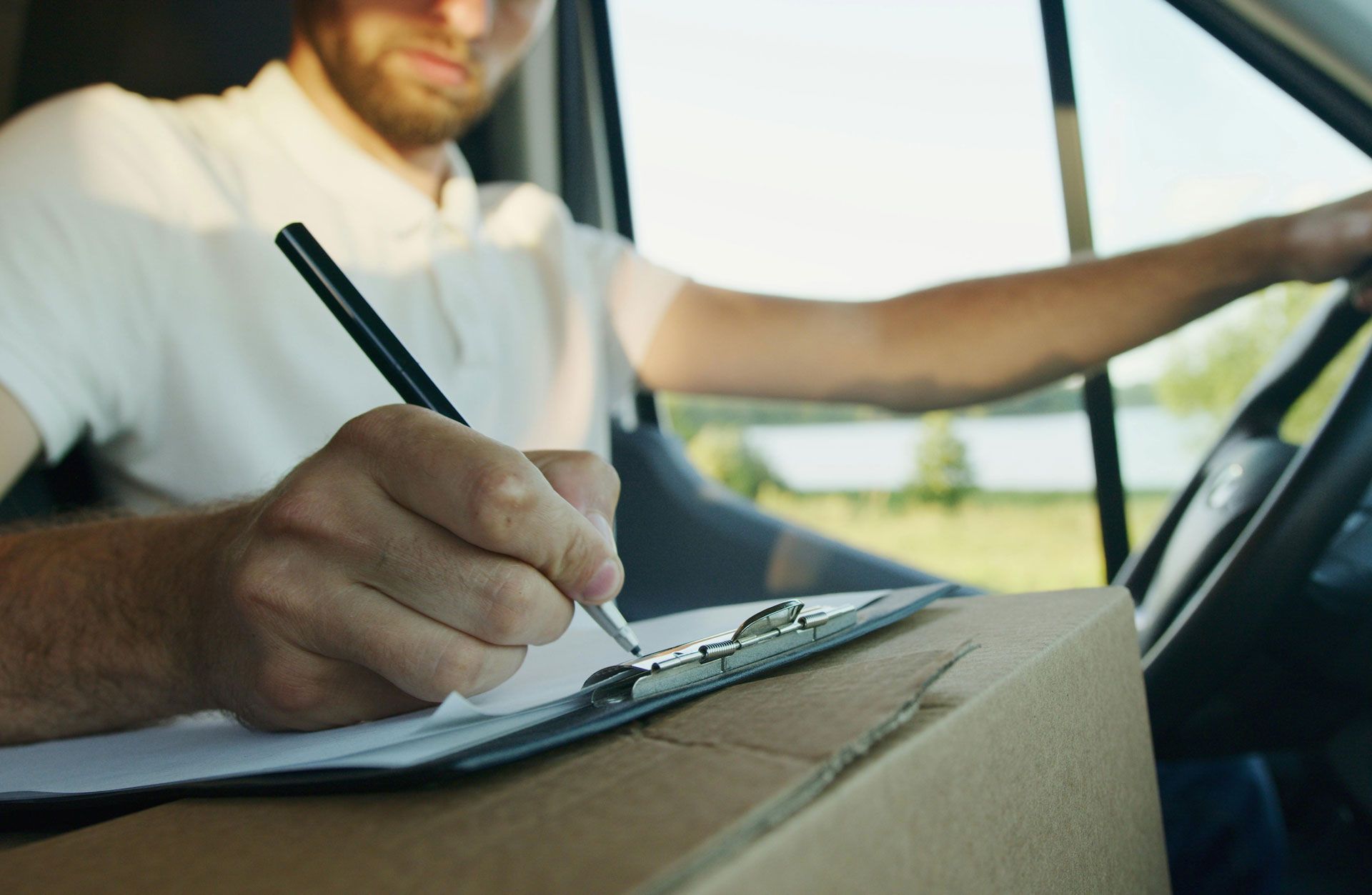 Delivery driver in van, writing on clipboard atop a cardboard box.