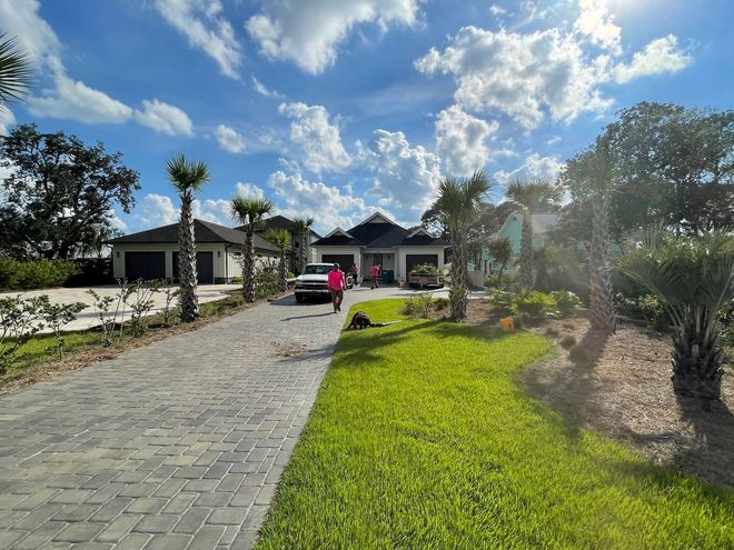 Gray brick driveway leads to a light-colored house on a sunny day, with lush green lawn and blue sky.