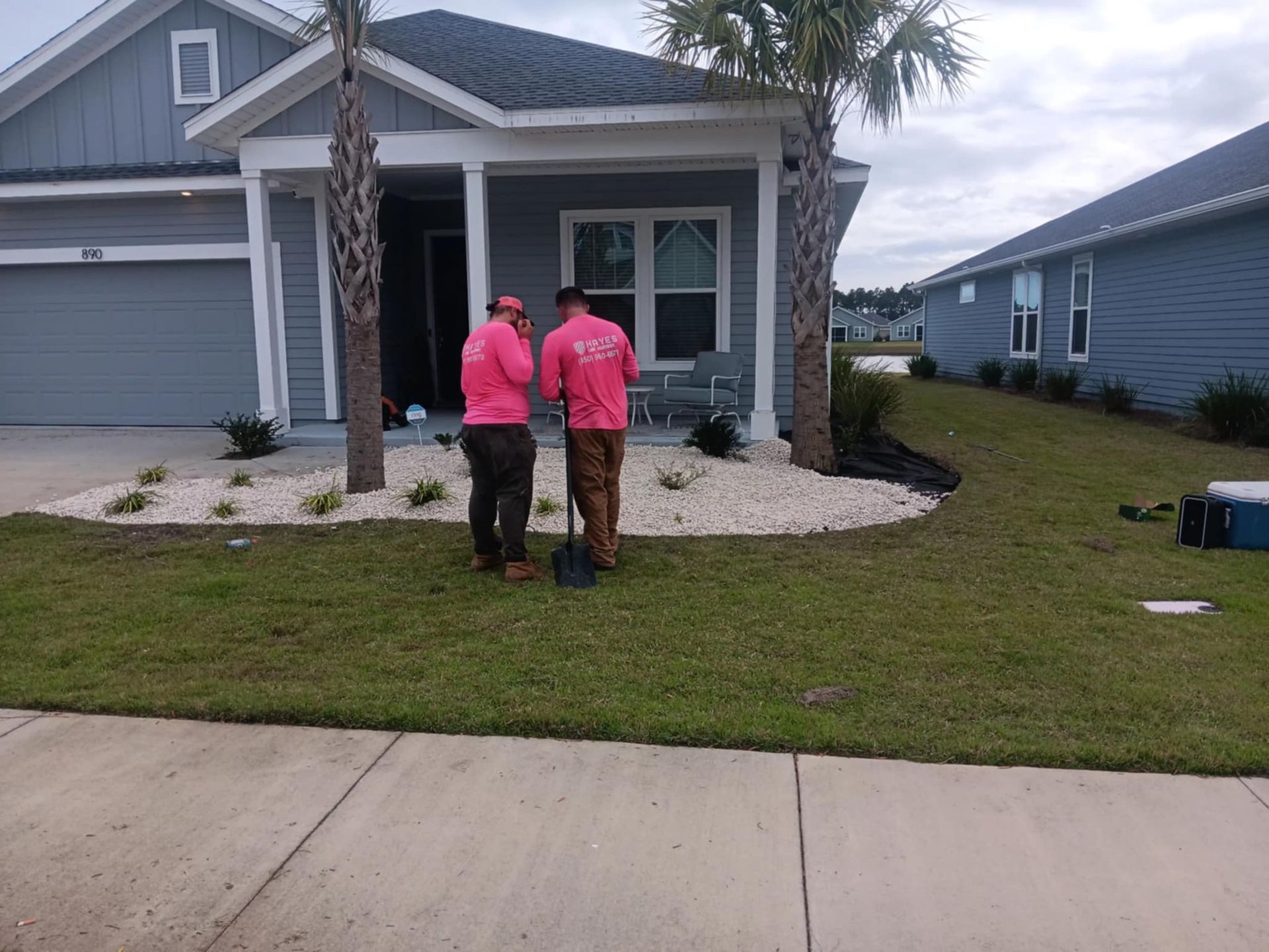 Two men in pink shirts landscaping a house with white rocks and palm trees.