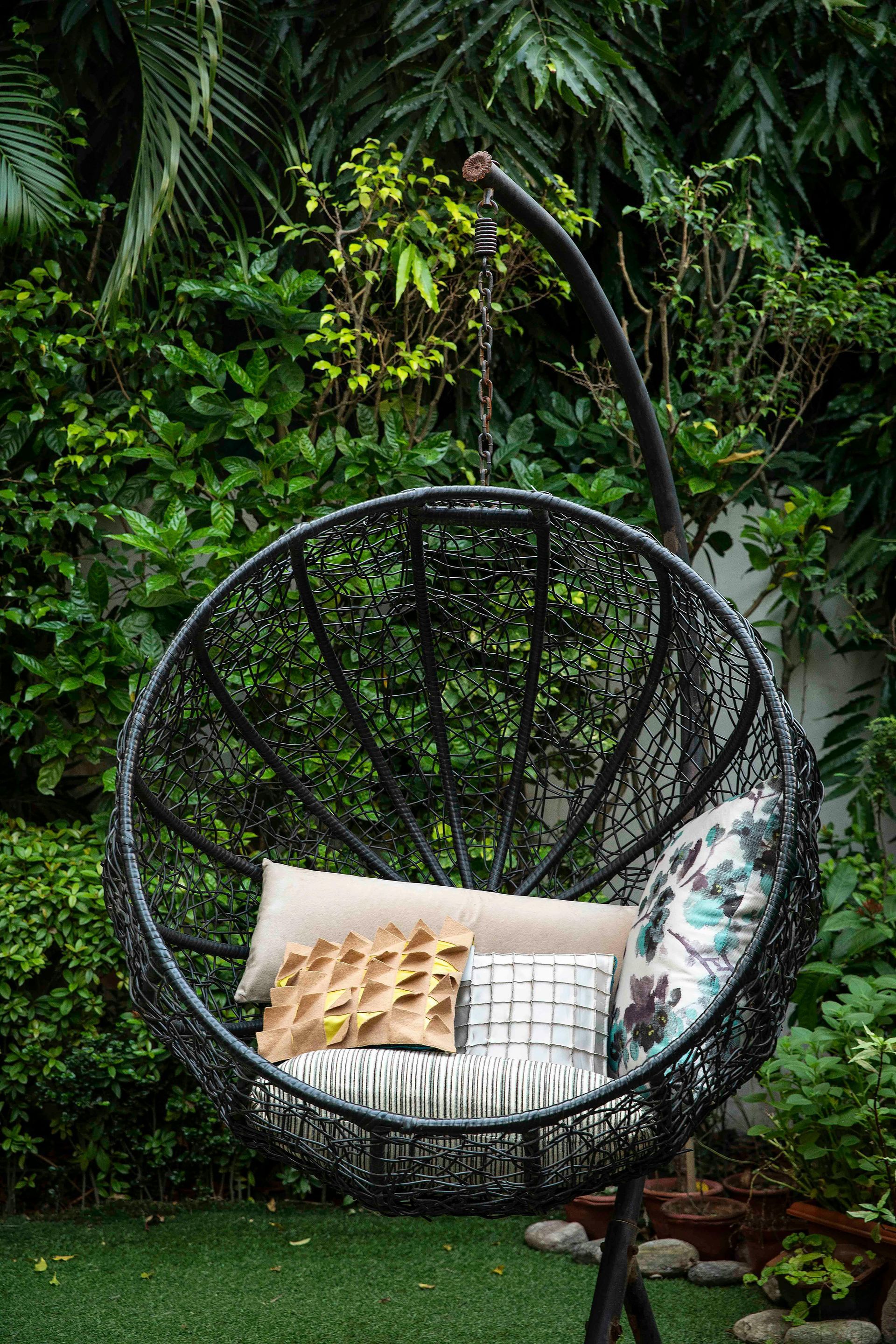Black wicker hanging chair with pillows in a lush green garden setting.