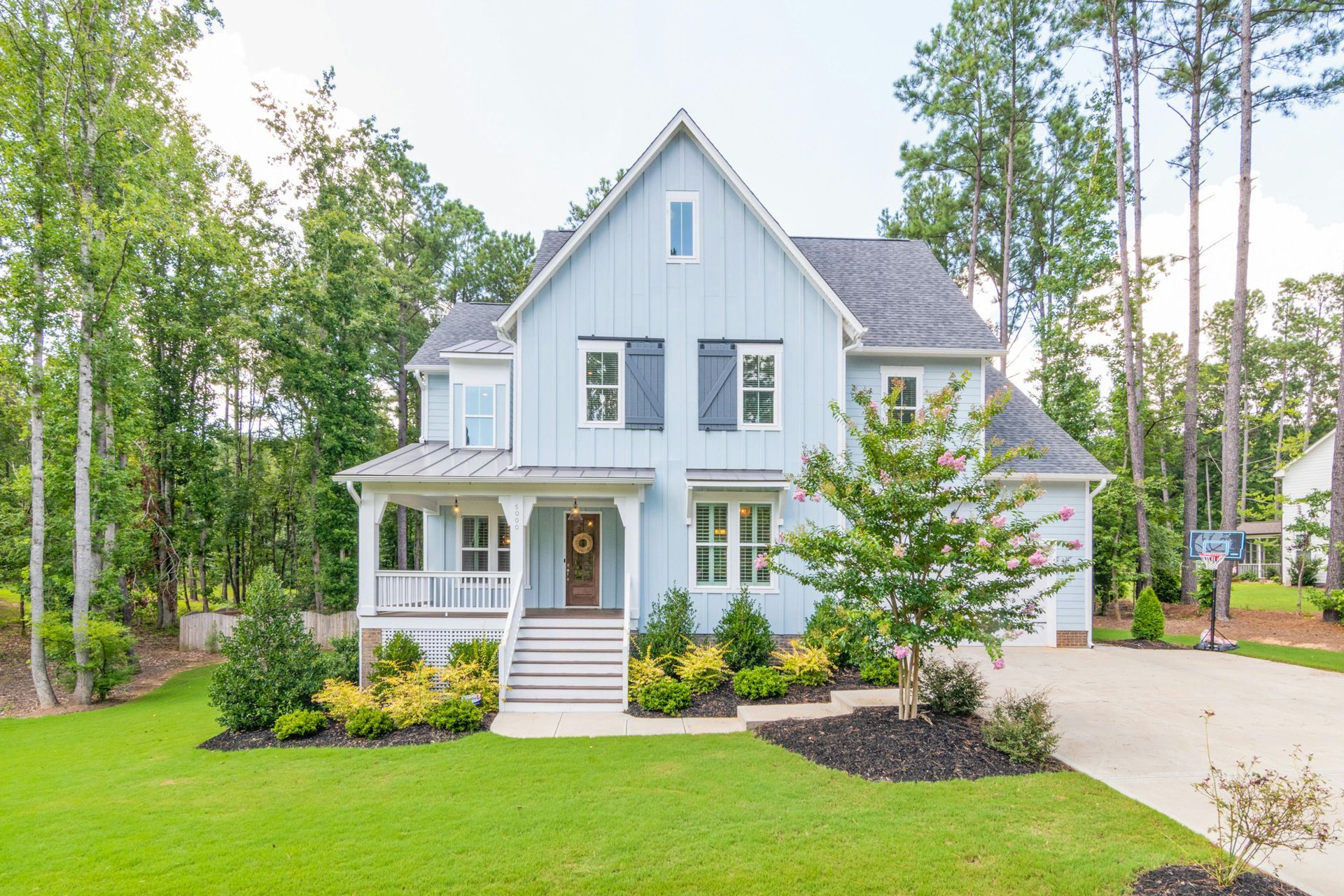 Blue two-story house with white trim, porch, and black shutters, set amongst trees on a green lawn.