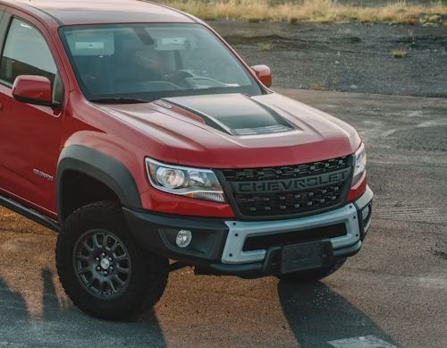 Red Chevrolet pickup truck with off-road tires and hood scoop parked on wet pavement.