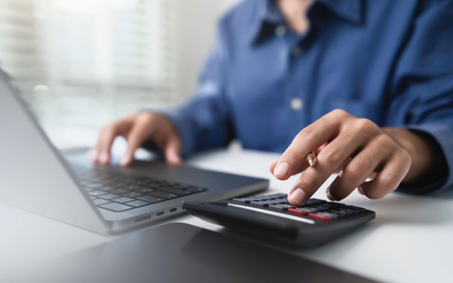 Person using a calculator and laptop, typing in a bright office.