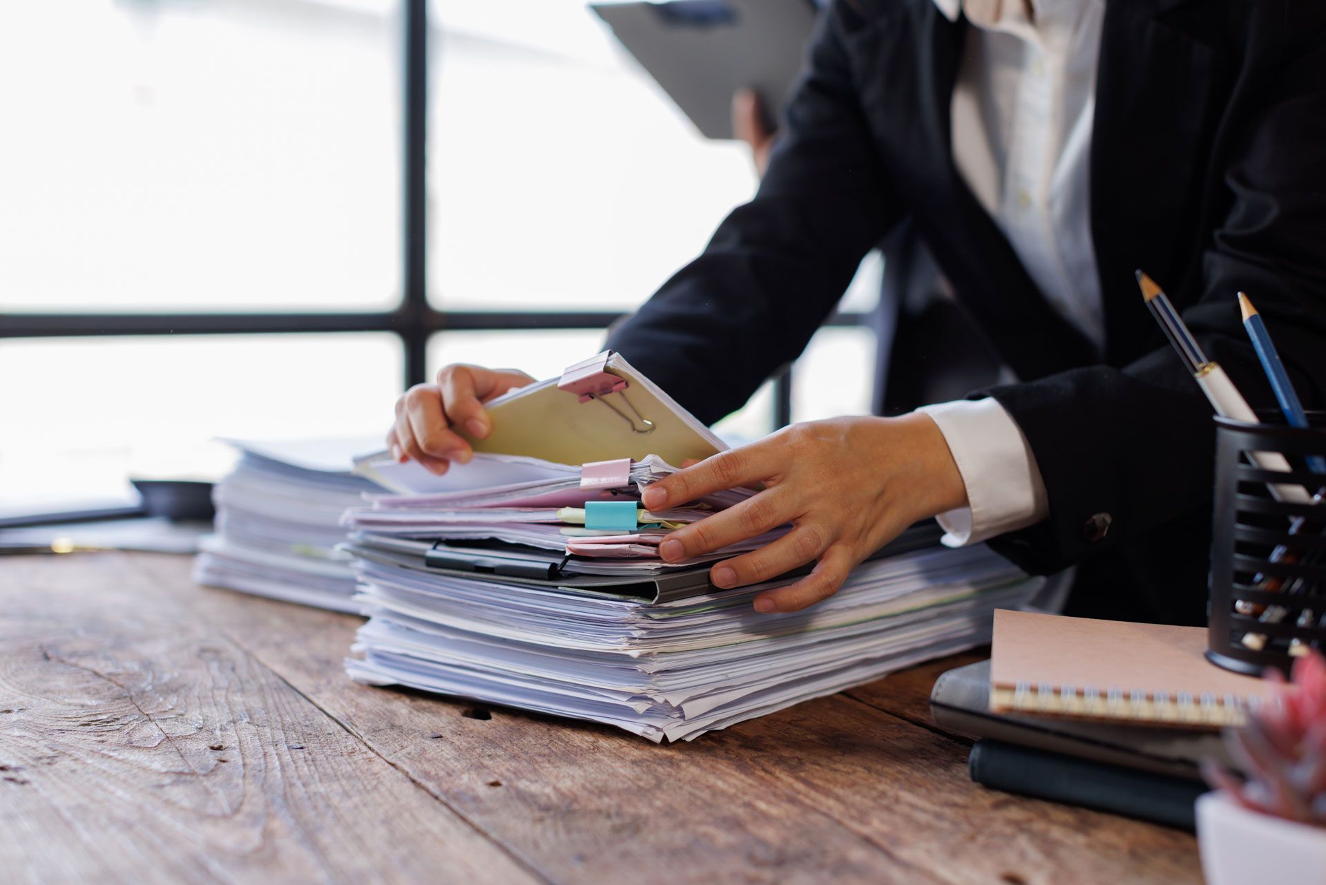 Person in blazer sorts through a stack of papers and files on a wooden desk.