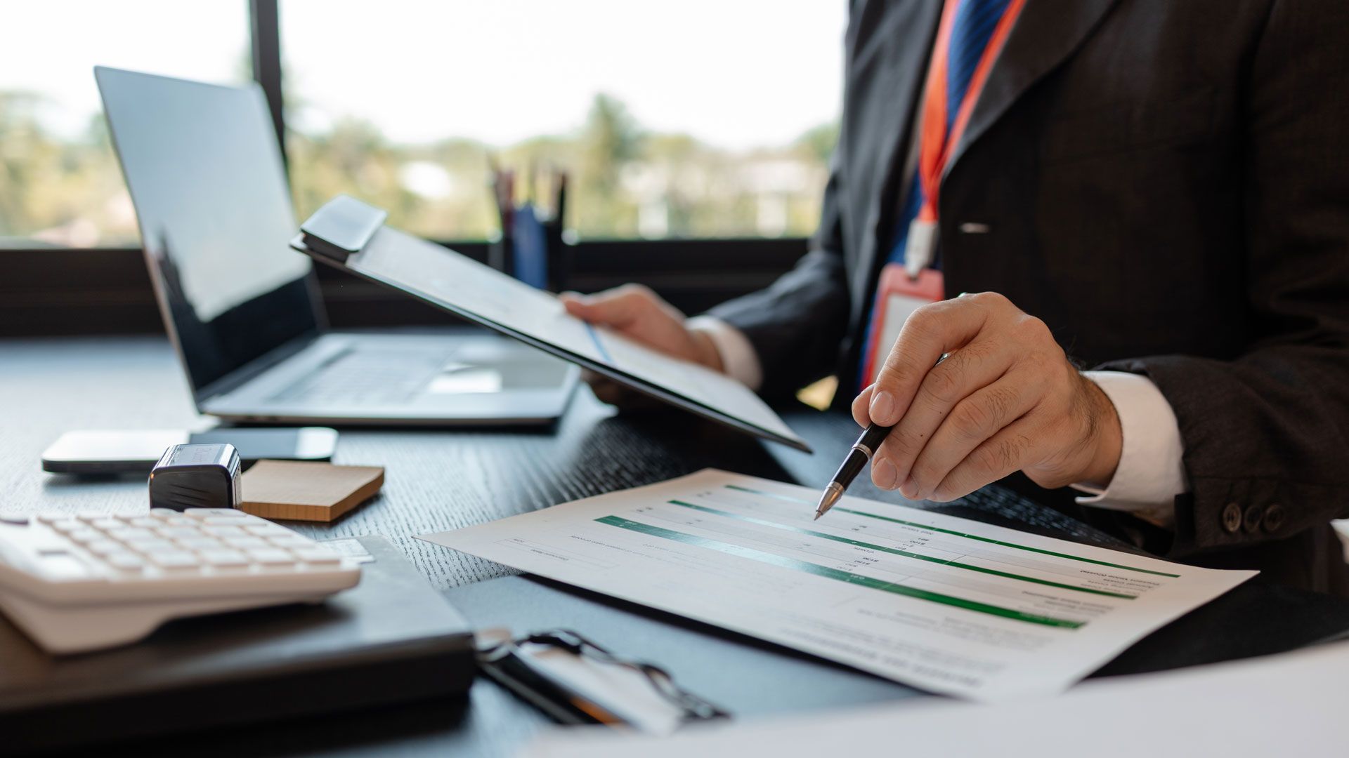 Person in suit reviewing documents at a desk, laptop, calculator, and window in the background.
