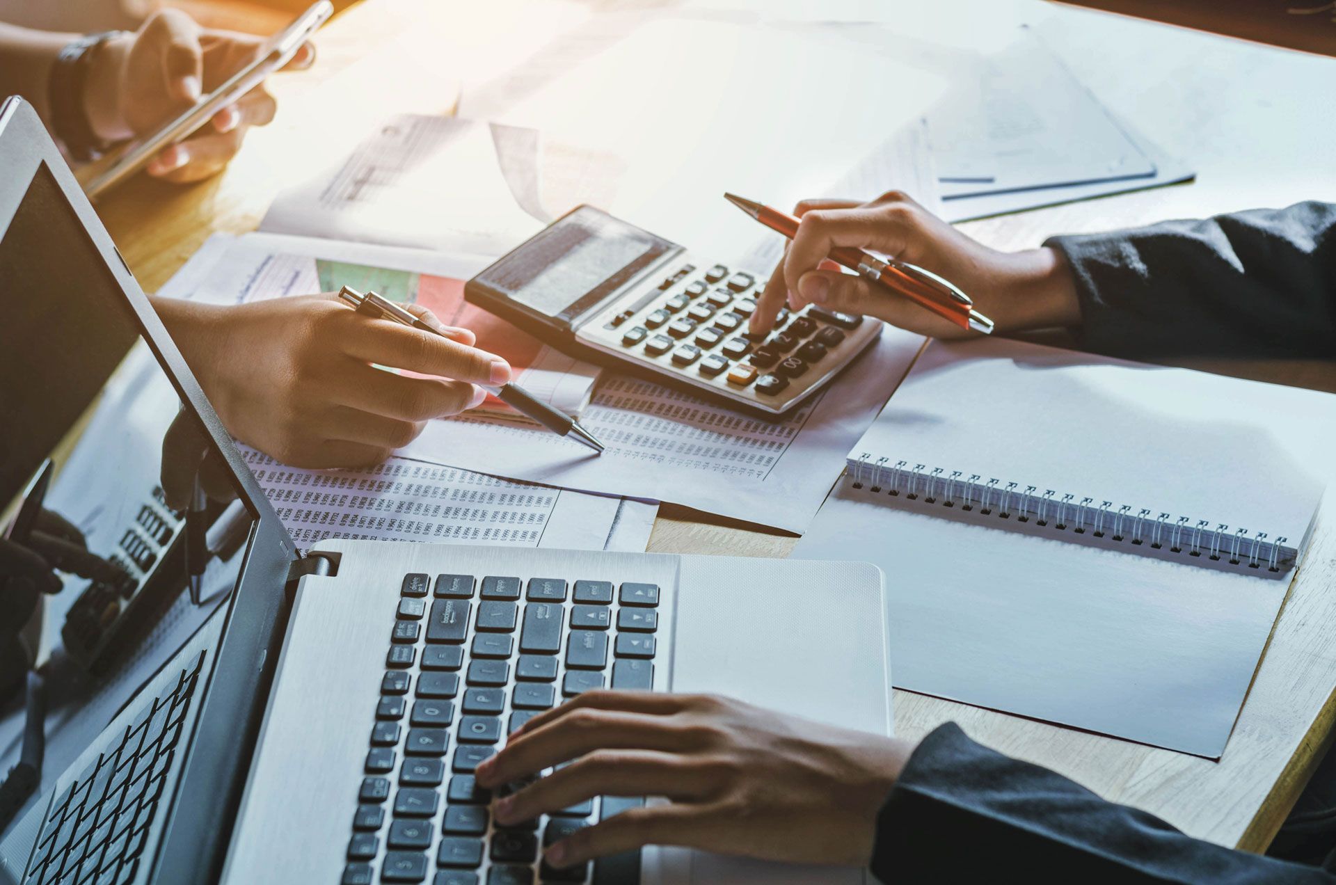 Hands using a laptop, calculator, phone, and papers on a desk; financial analysis in progress.