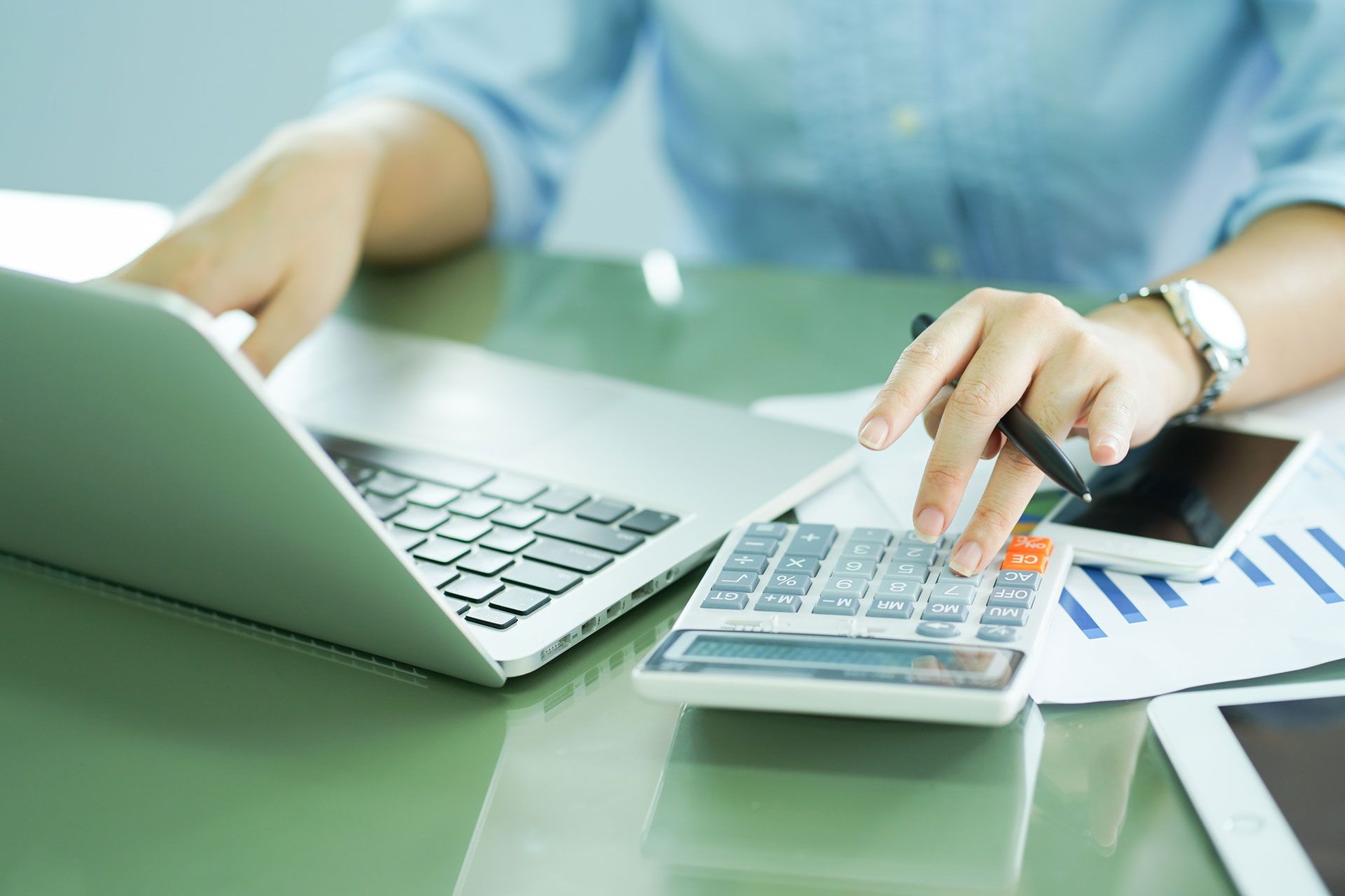 Person using a calculator, laptop, and smartphone on a green desk with financial charts.