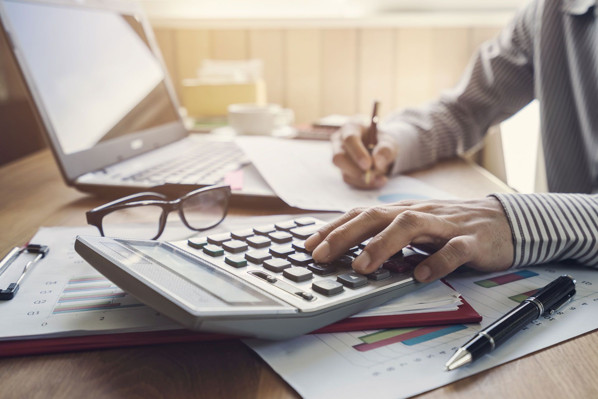 Person calculating with a calculator at a desk, laptop, pen, glasses, and documents are present.