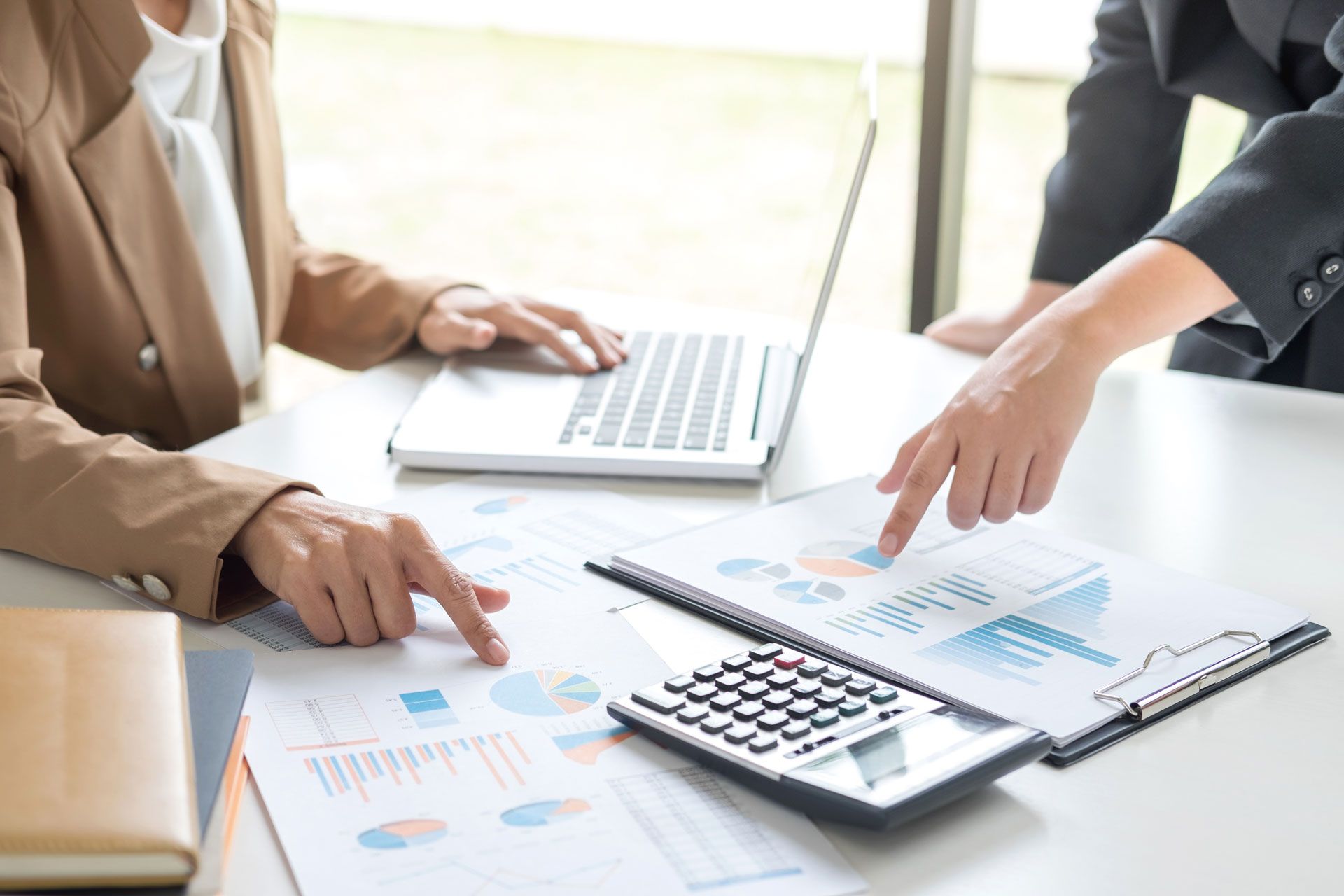 Two people at a desk review data charts, pointing at the graphs and using a calculator, laptop present.