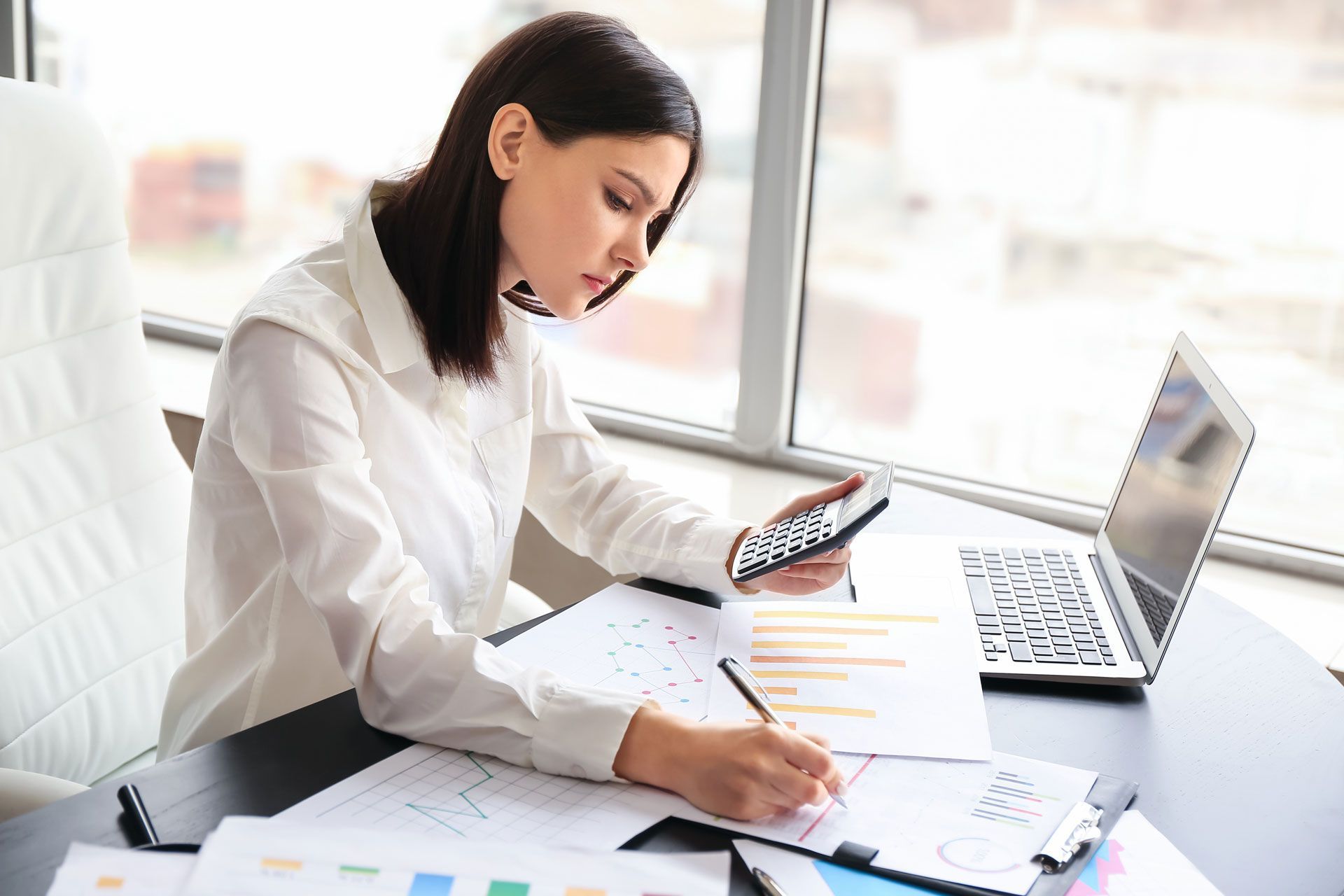 Woman using calculator, laptop, and documents at desk by window.