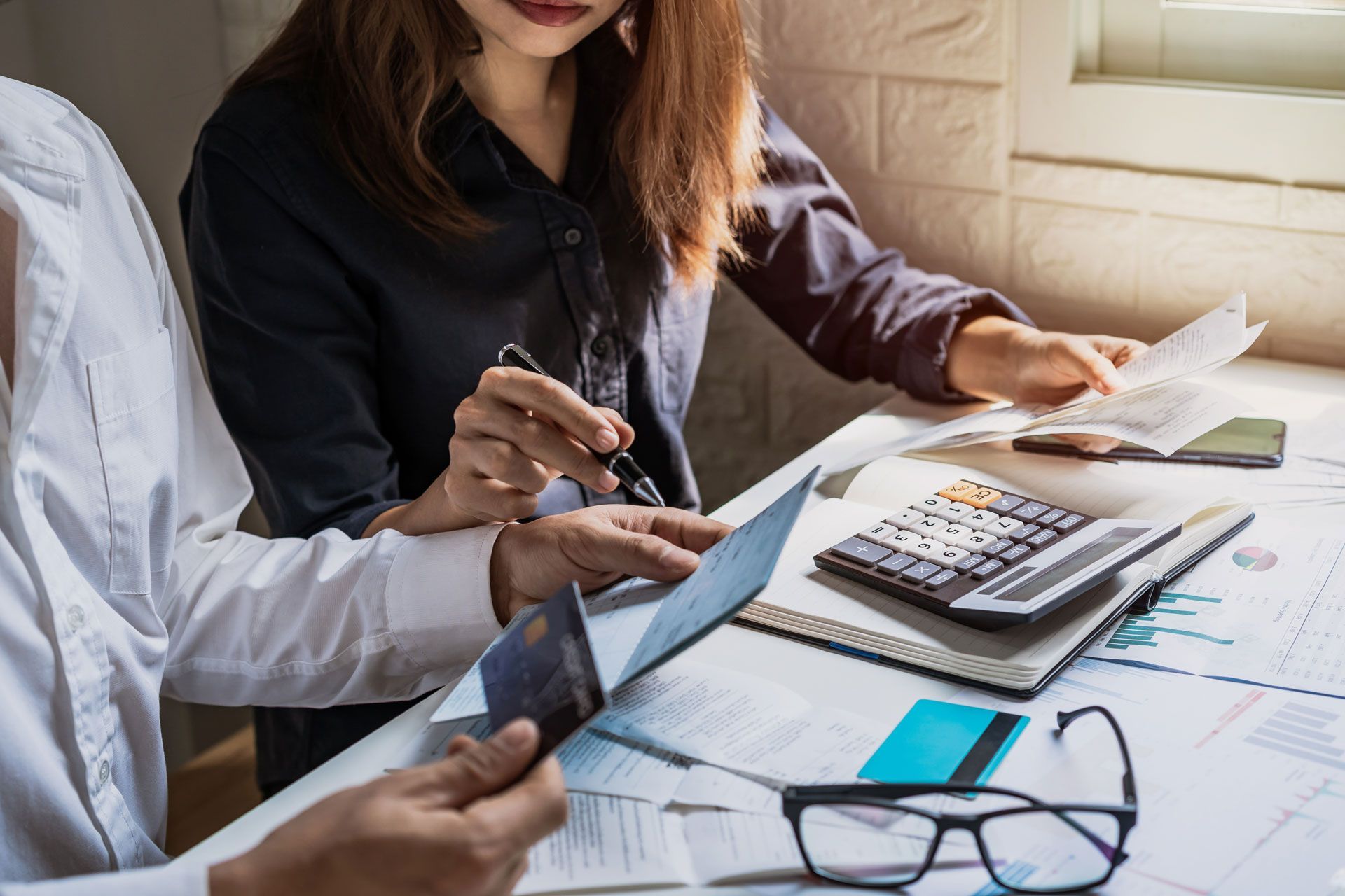 Two people reviewing financial documents with a calculator, credit cards, and glasses on a desk.