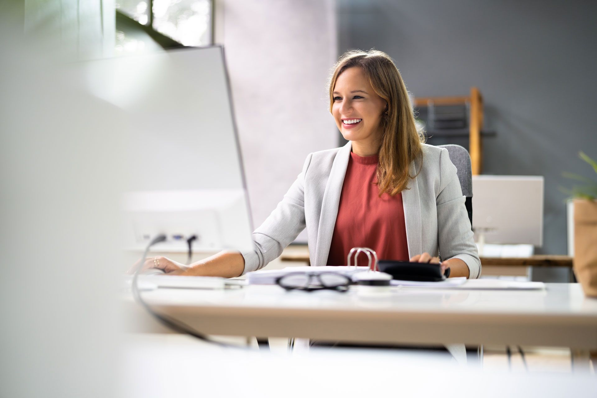 Woman smiling while working at a computer in a modern office, wearing a gray blazer and red shirt.
