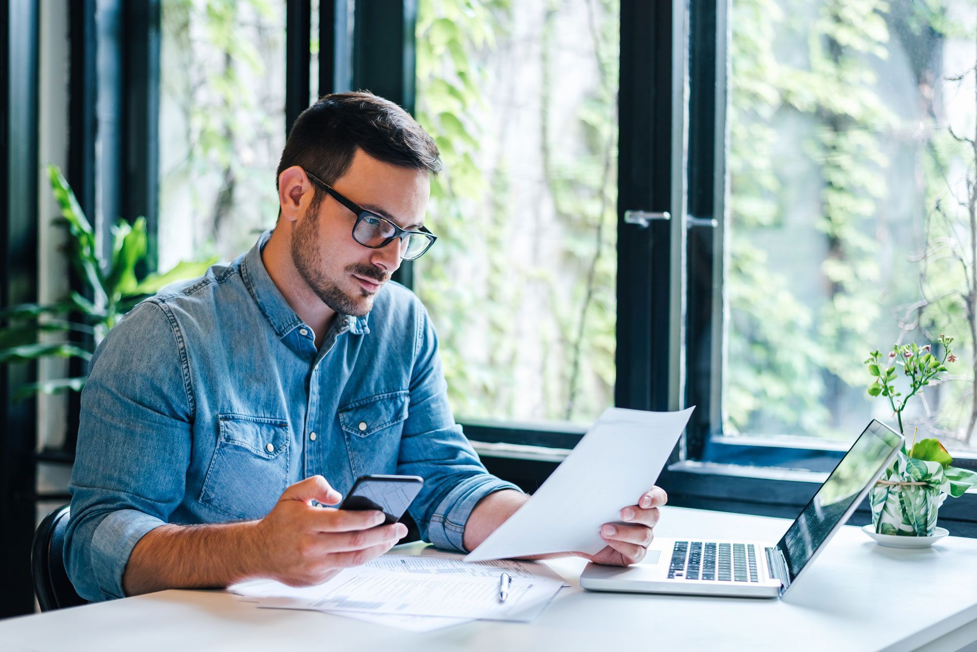 Man with glasses in blue shirt, using phone, holding paper, working on a laptop at a desk by a window.