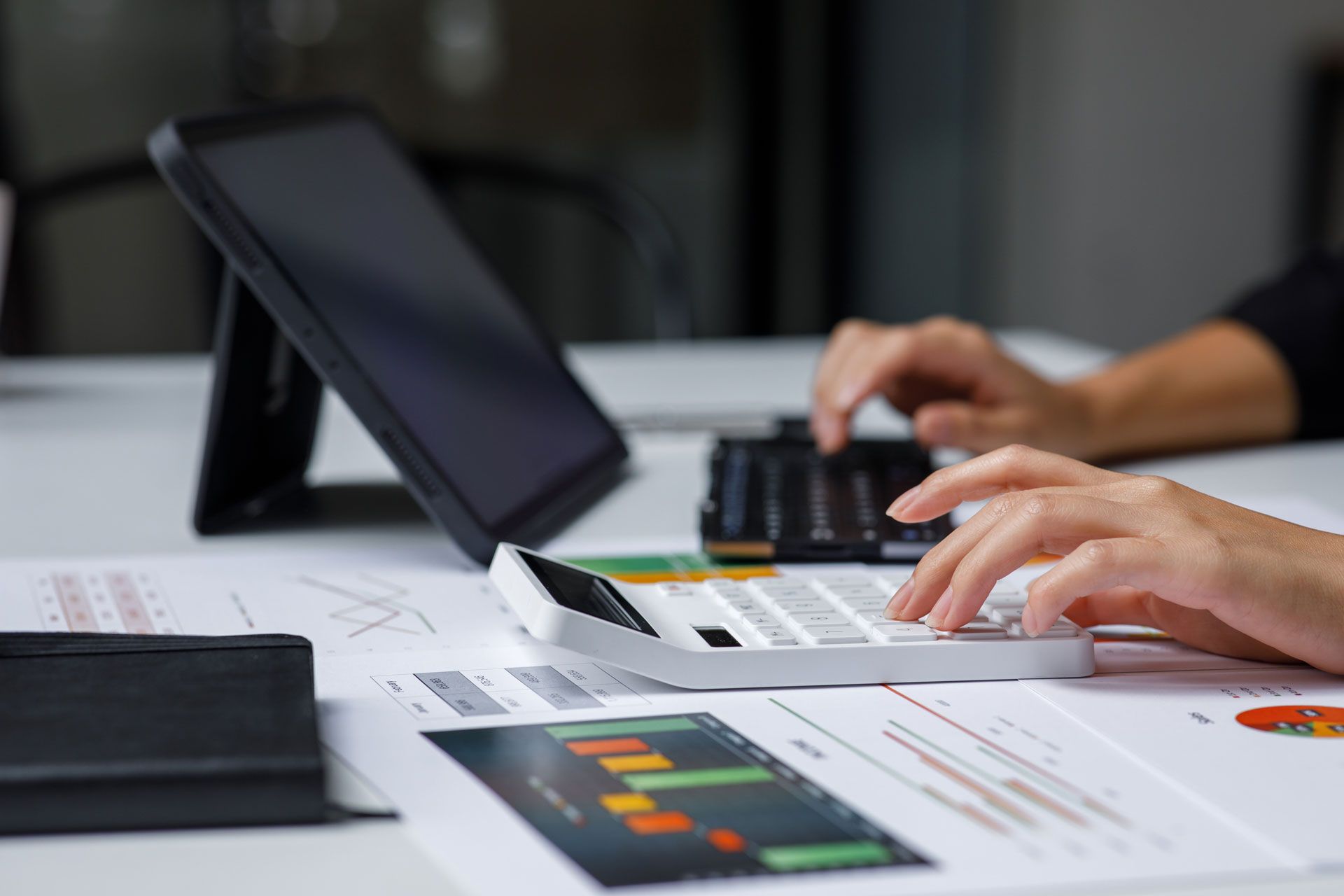 Hands using calculator over financial documents, tablet visible in background.