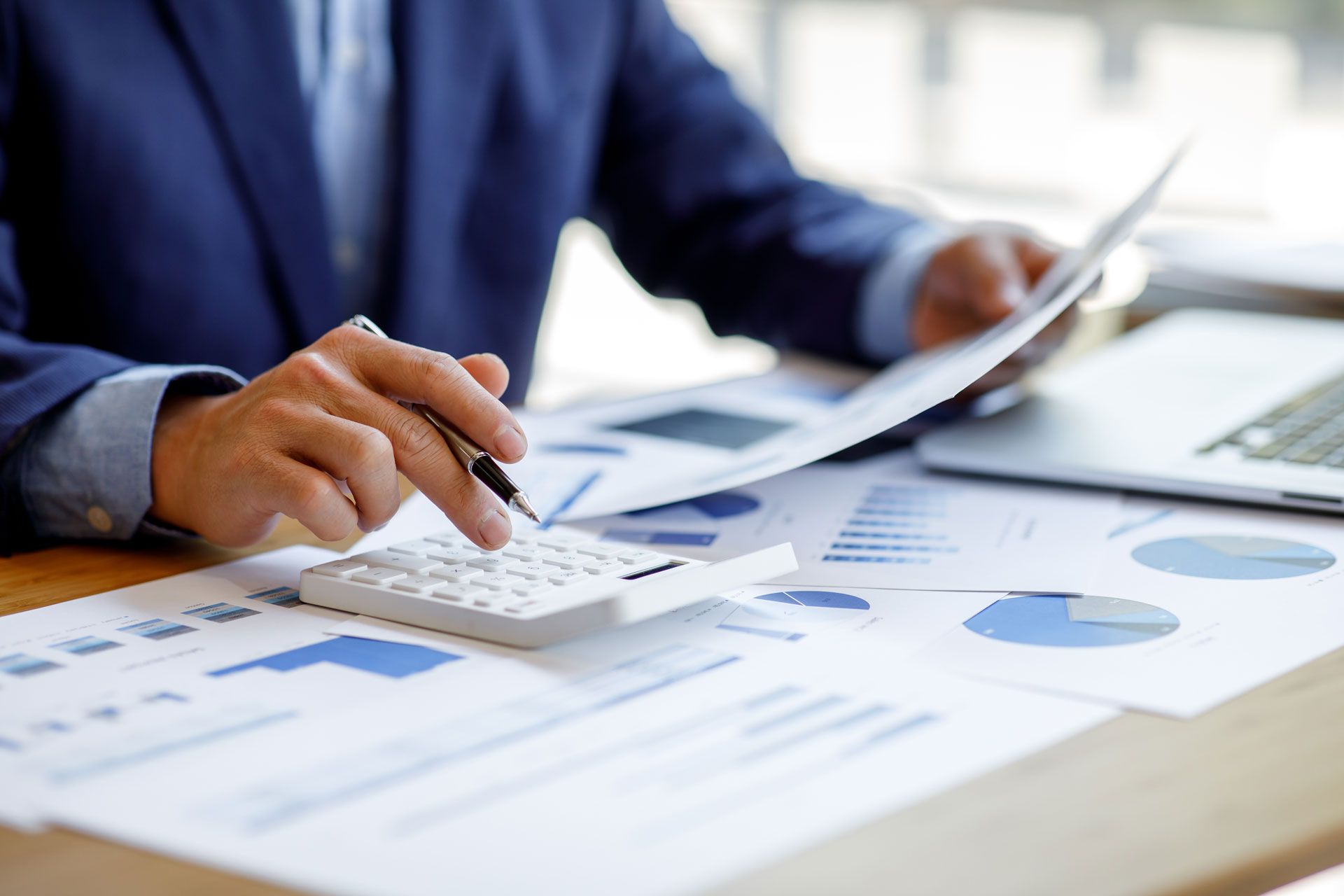 Person in blue suit calculating finances with papers, calculator, and laptop on desk.
