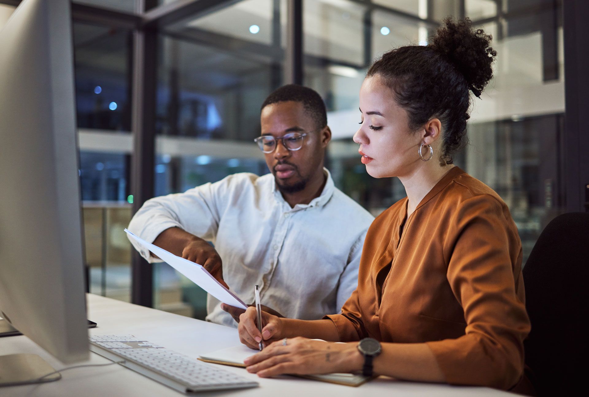 Two people reviewing documents at a desk in an office. One points, the other writes.