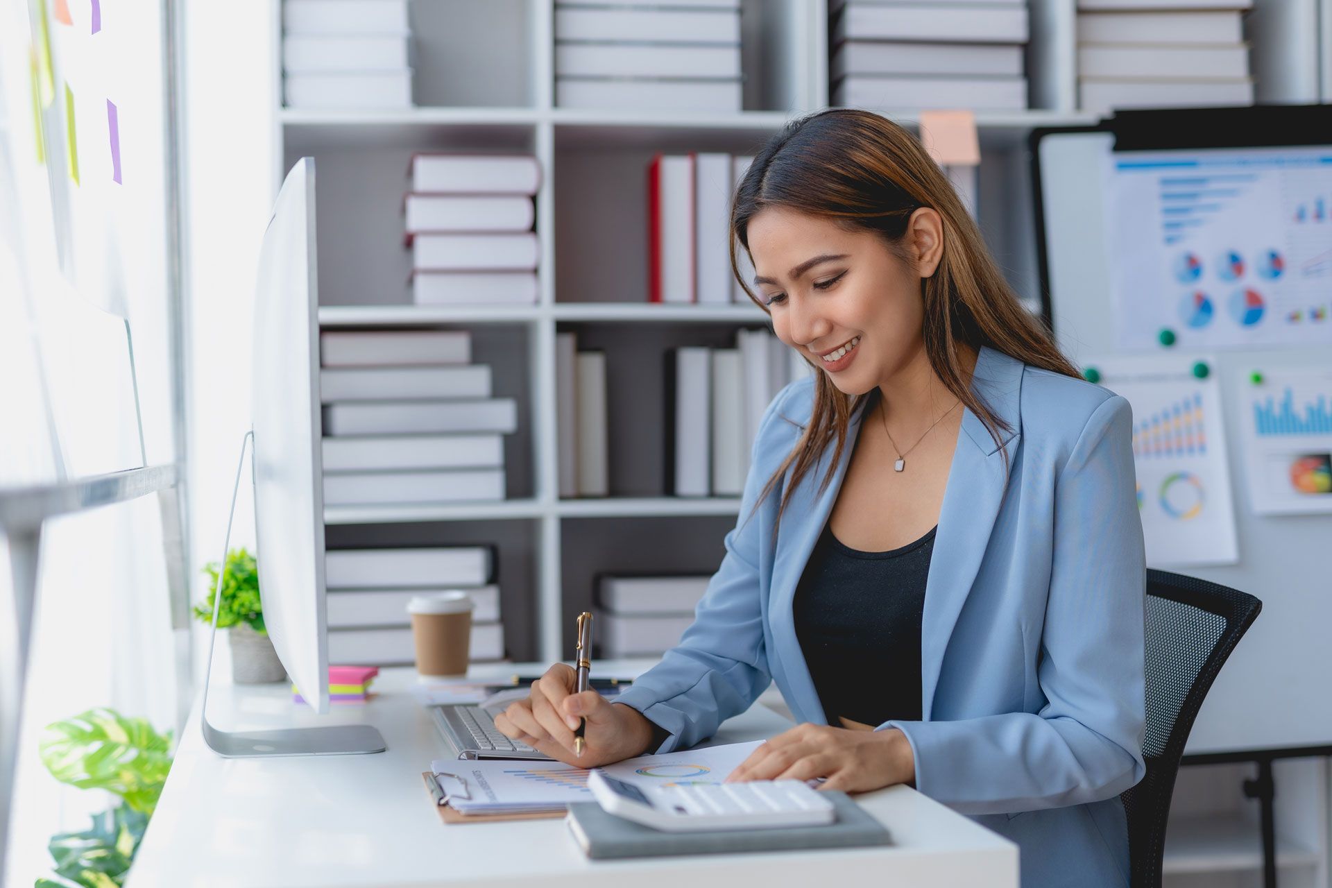 Woman in blue blazer works at desk, writing on paper; white bookshelf in background.